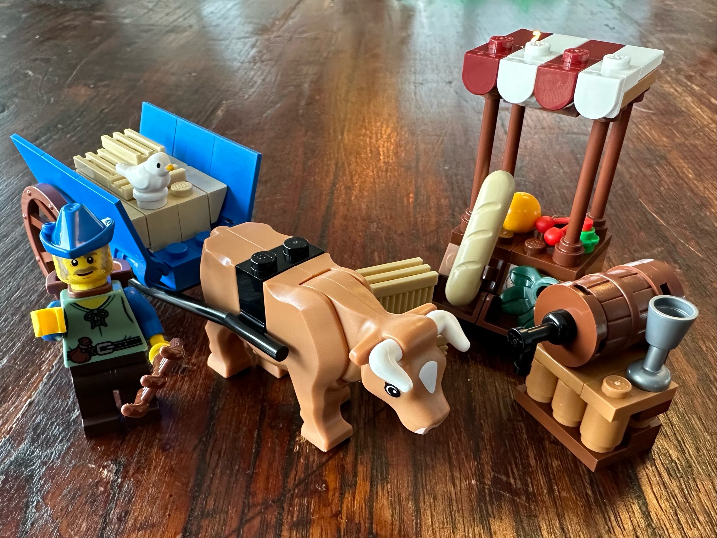LEGO peasant holding a pitchfork, a cow drawing a cart filled with hay, a small market stocked with bread and produce, and a small table holding a barrel of booze and a goblet.