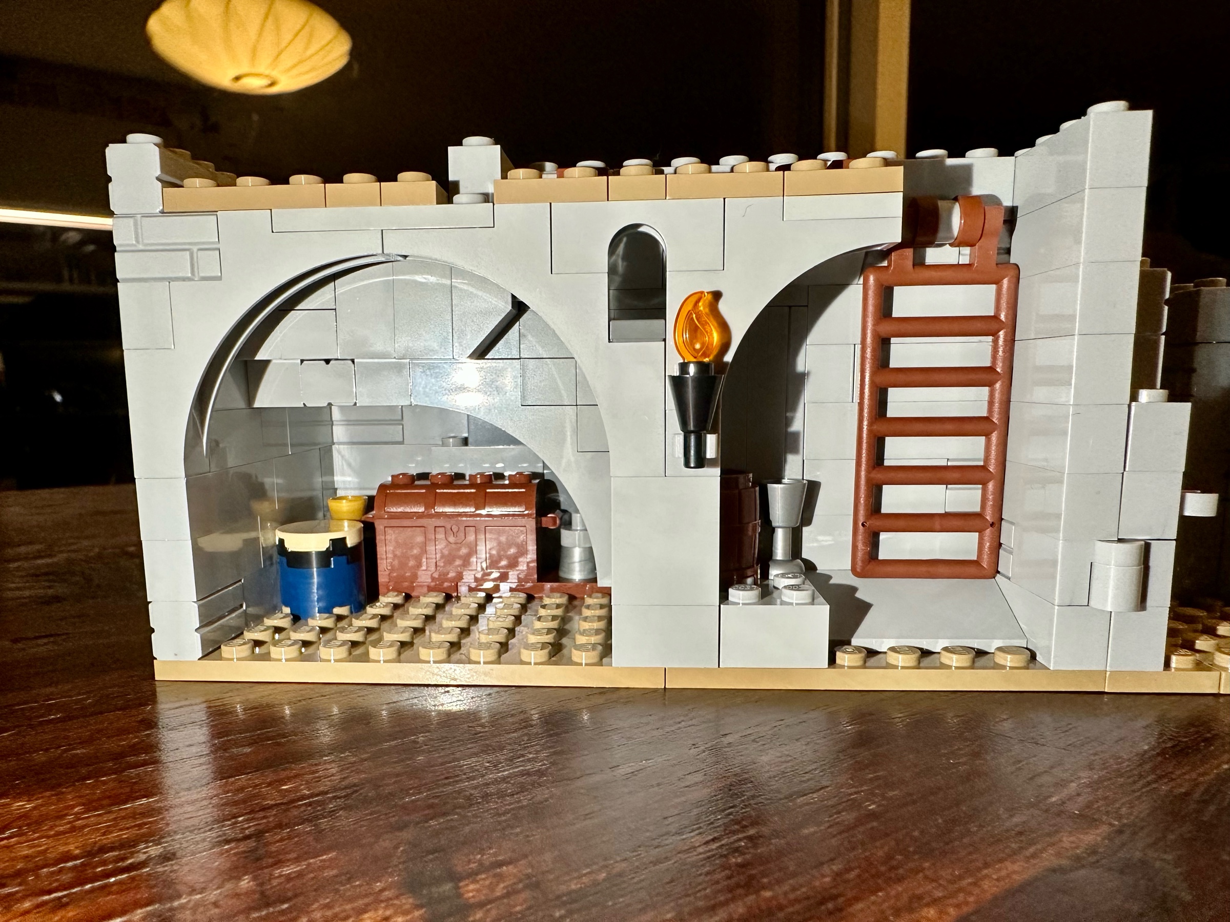 A LEGO dungeon. A treasure vault holding a chest, a goblet, a drum, and a bell is on the left. A wooden ladder and a flaming torch are on the right.