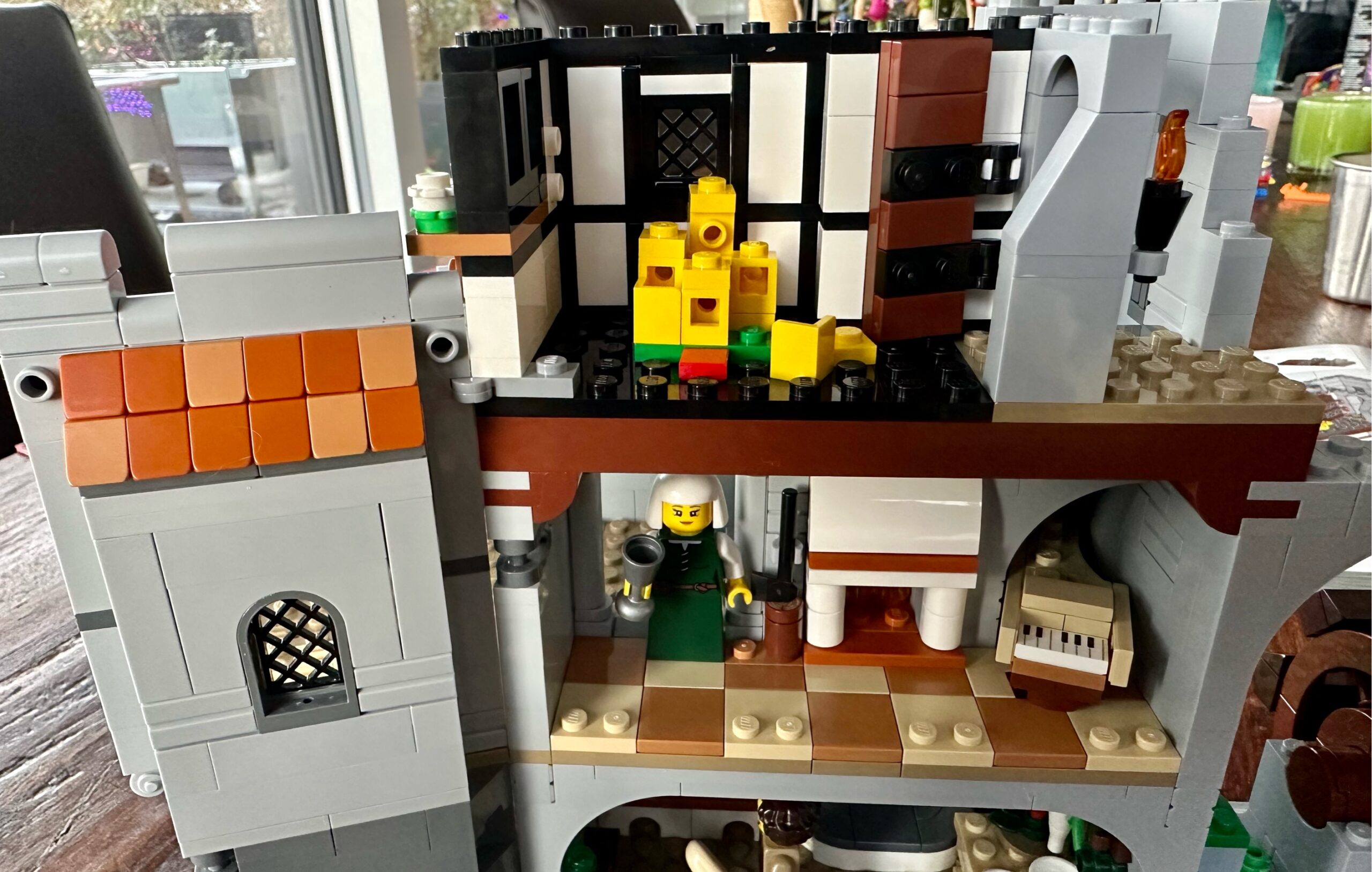 Interior view of LEGO castle with a bedroom with a toy castle set above a music room. To the left is a wall with a window and a bit of tiled roof.
