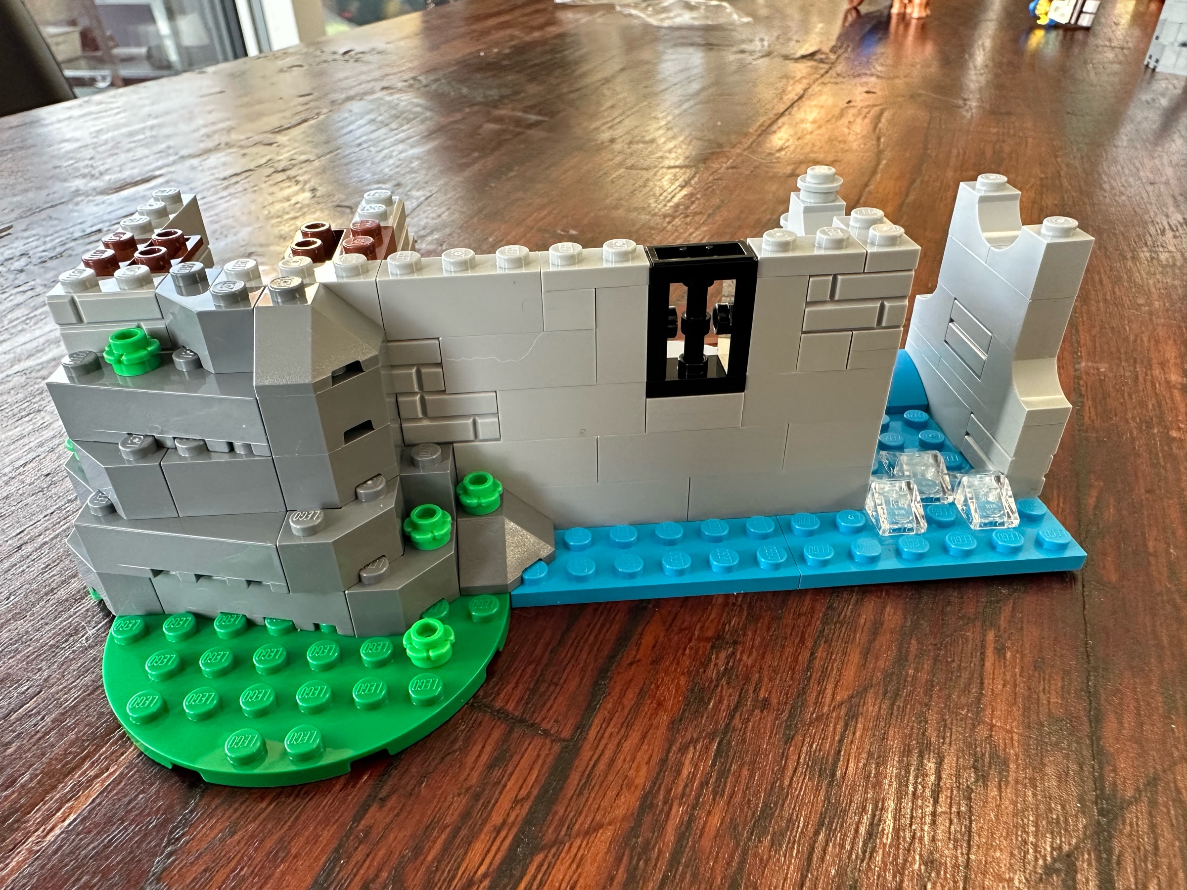 Exterior view of LEGO castle showing gray stone walls, a bit of greenery on the left, and a flowing stream with a few rapids on the right.