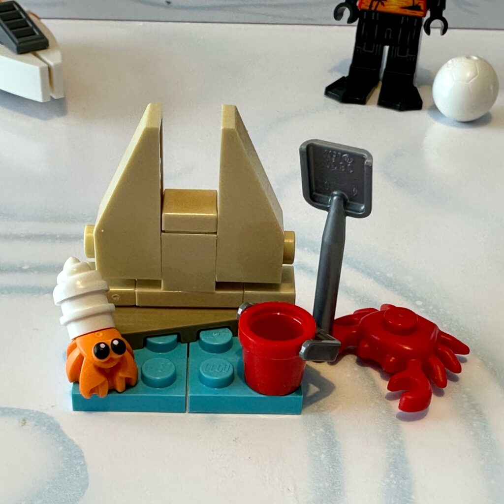 LEGO micro scale build of a sand castle on the water’s edge together with a bucket, a cute hermit crab, and red crab holding a shovel in its claw.