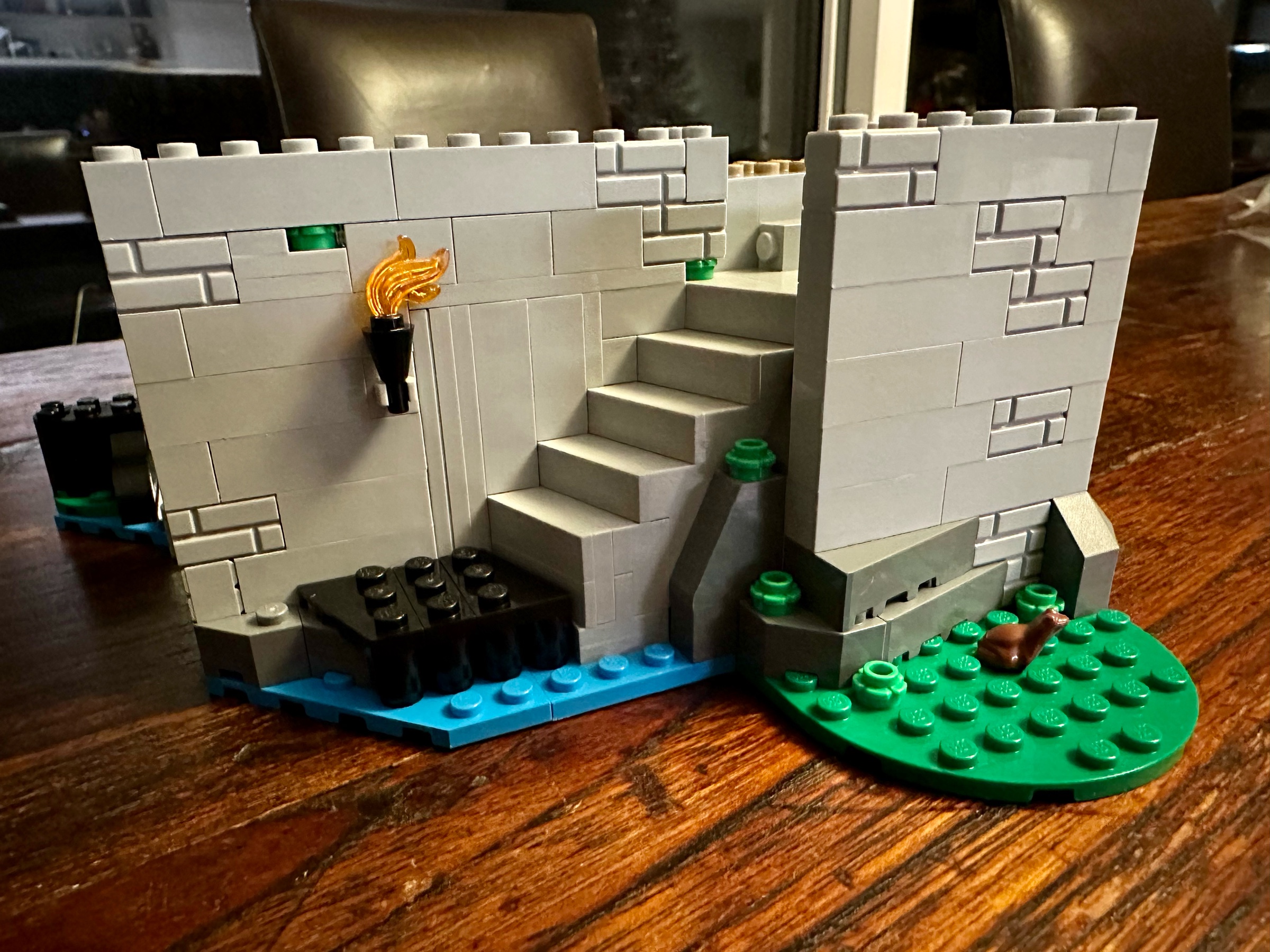 Exterior view of LEGO castle with a staircase leading up from a small dock. The castle wall by the stairs bears a flaming torch. A brown frog sits on the grass nearby.