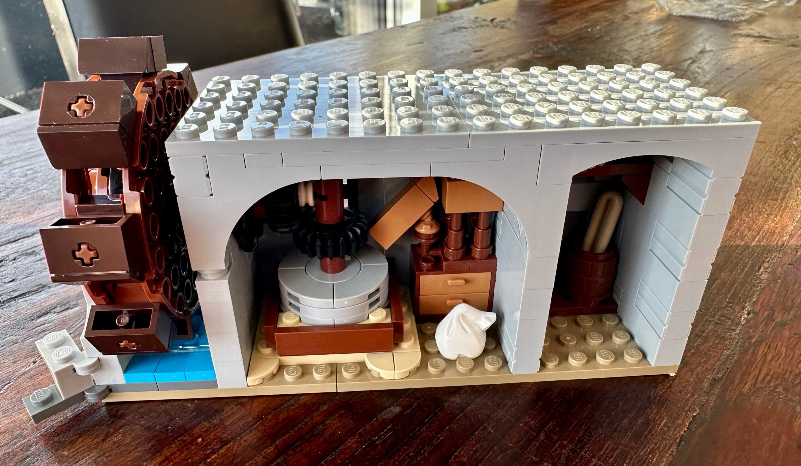 Interior view of LEGO castle section with a water wheel connected to a millstone. A storage system and hopper feeds grain to the millstone. A sack (containing flour?) sits on the floor. A storage room has a baguette stuffed into a barrel and a shelf with a bird's nest.