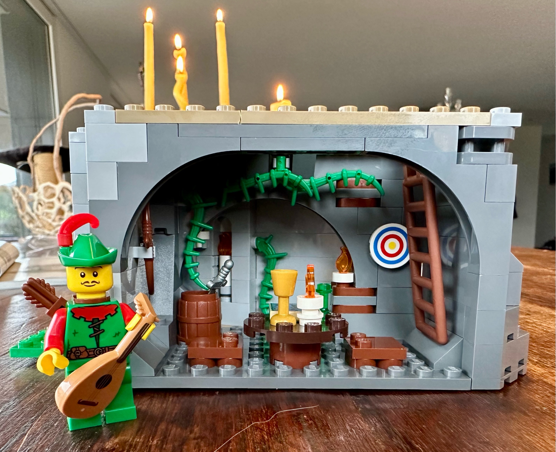 Small LEGO cave with a table, stools, candles, goblet and a map. An archery target hangs on the wall and there are vines along the ceiling.