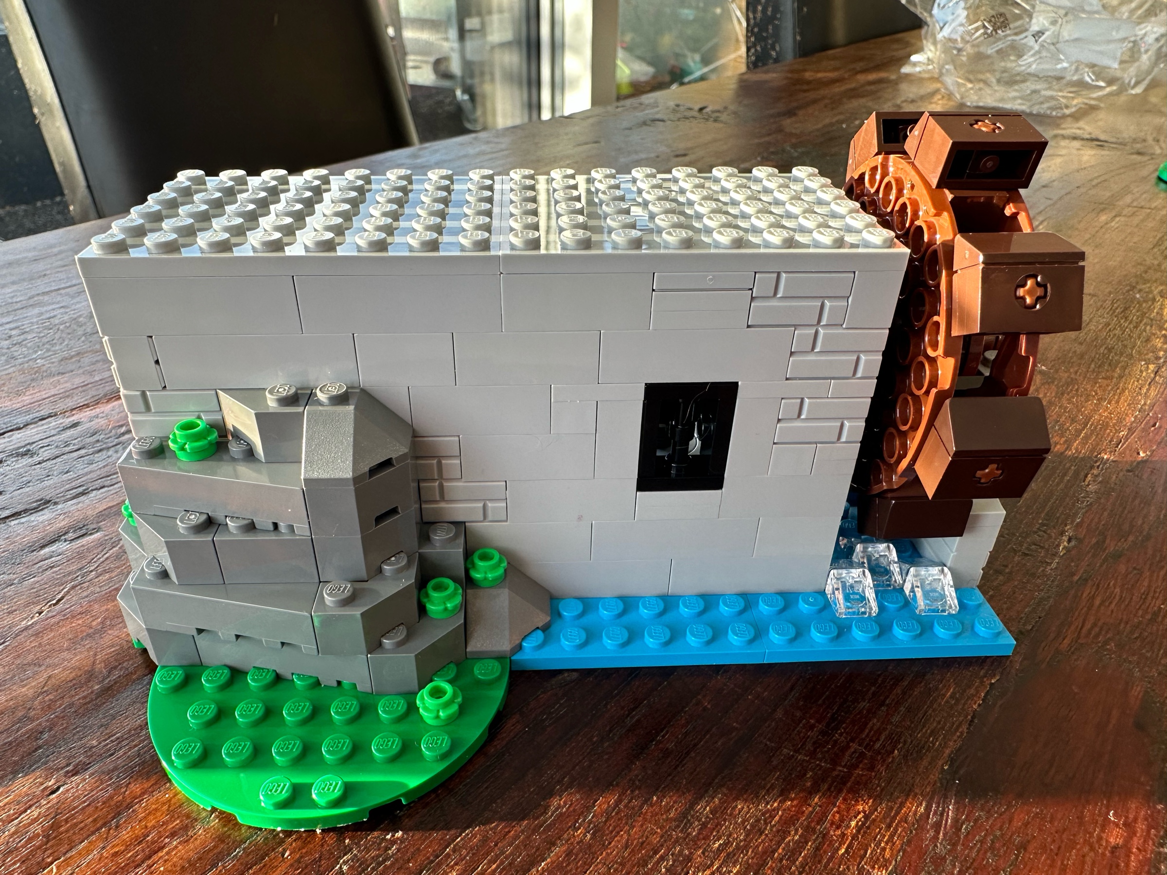 Exterior view of LEGO castle section showing a water wheel over a flowing stream on the right, a single low barred window in the center, and a rocky outcropping on the left.