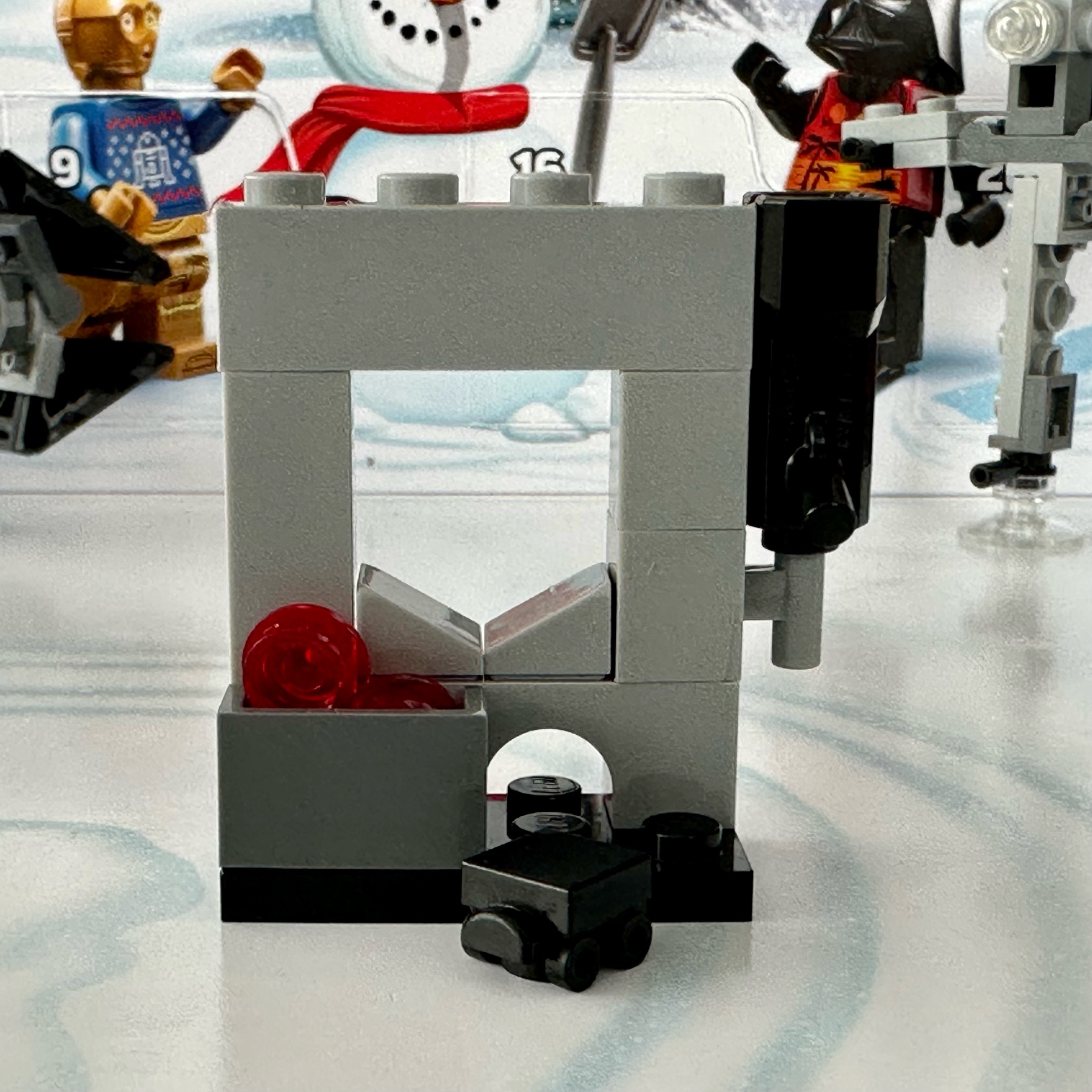 LEGO build of a small section of wall with a window and mouse hole in it. A gun hangs on the wall and a small crate of ammo sits in front. A mouse droid has rolled out of the hole.