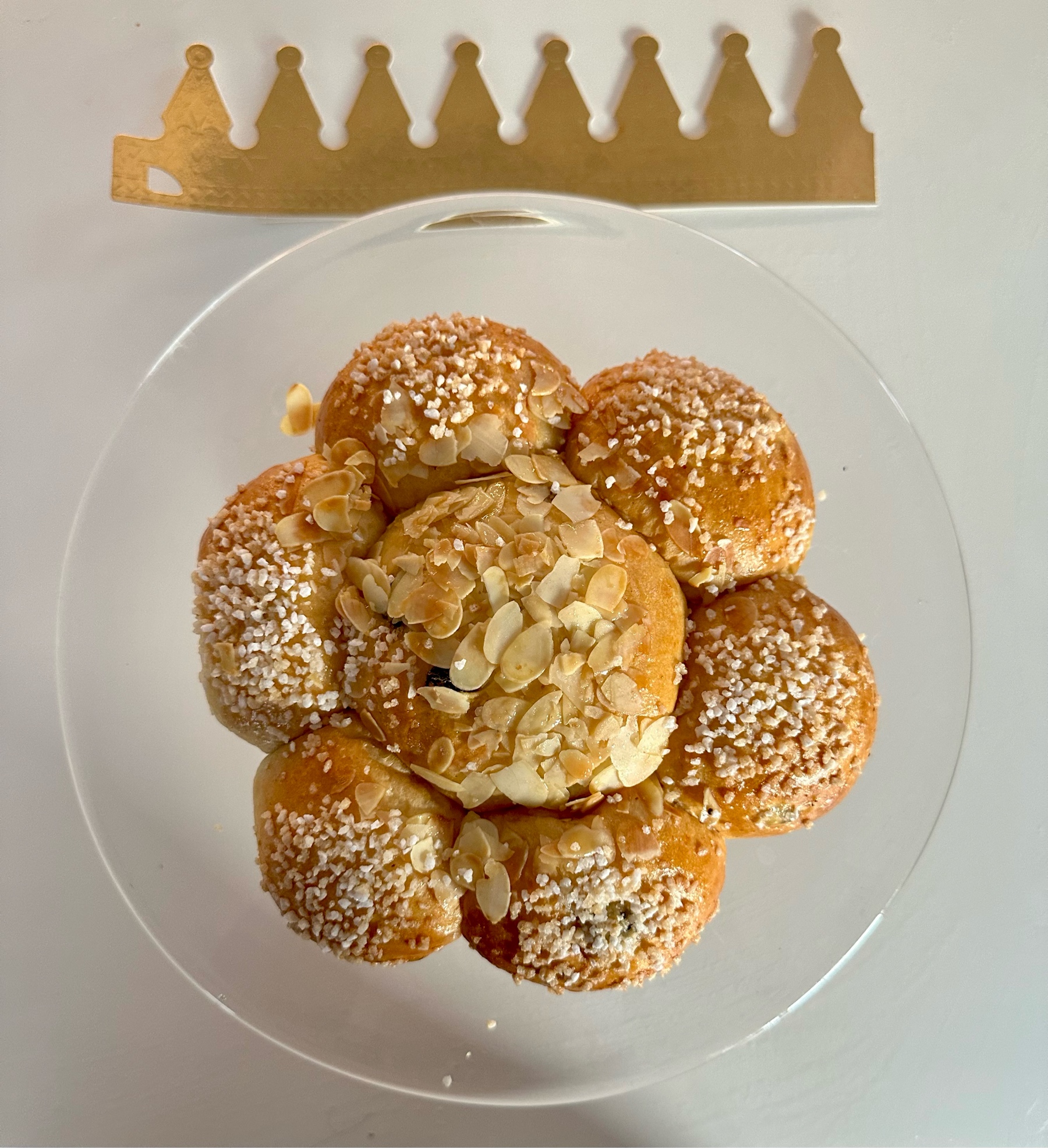Sweet roll covered in almond slivers and coarse sugar with 6 rolls around a larger center. A gold paper crown is at the top.