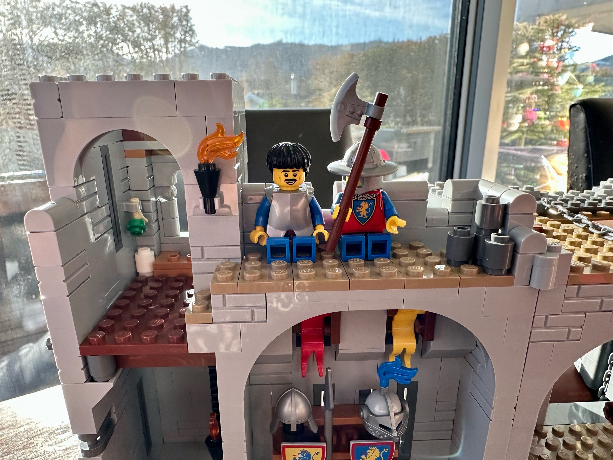 LEGO castle parapet with a latrine on the left. Two guards sit on the parapet with their backs to the wall. A pile of stones to drop through the murder holes is on the right.
