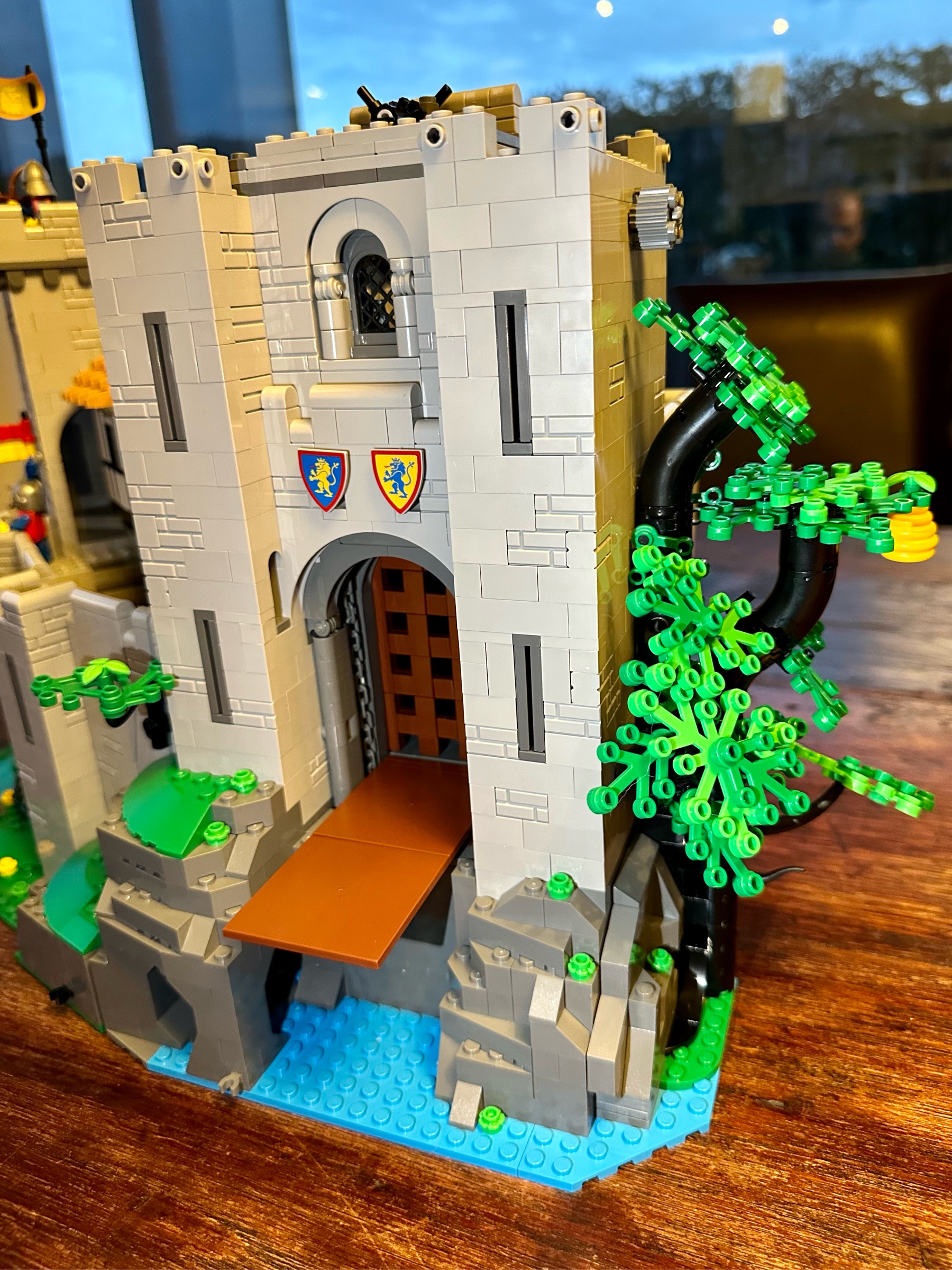 Exterior view of LEGO castle with drawbridge lowered revealing a closed portcullis behind it. A tree on the right side had lush green foliage and a yellow beehive.