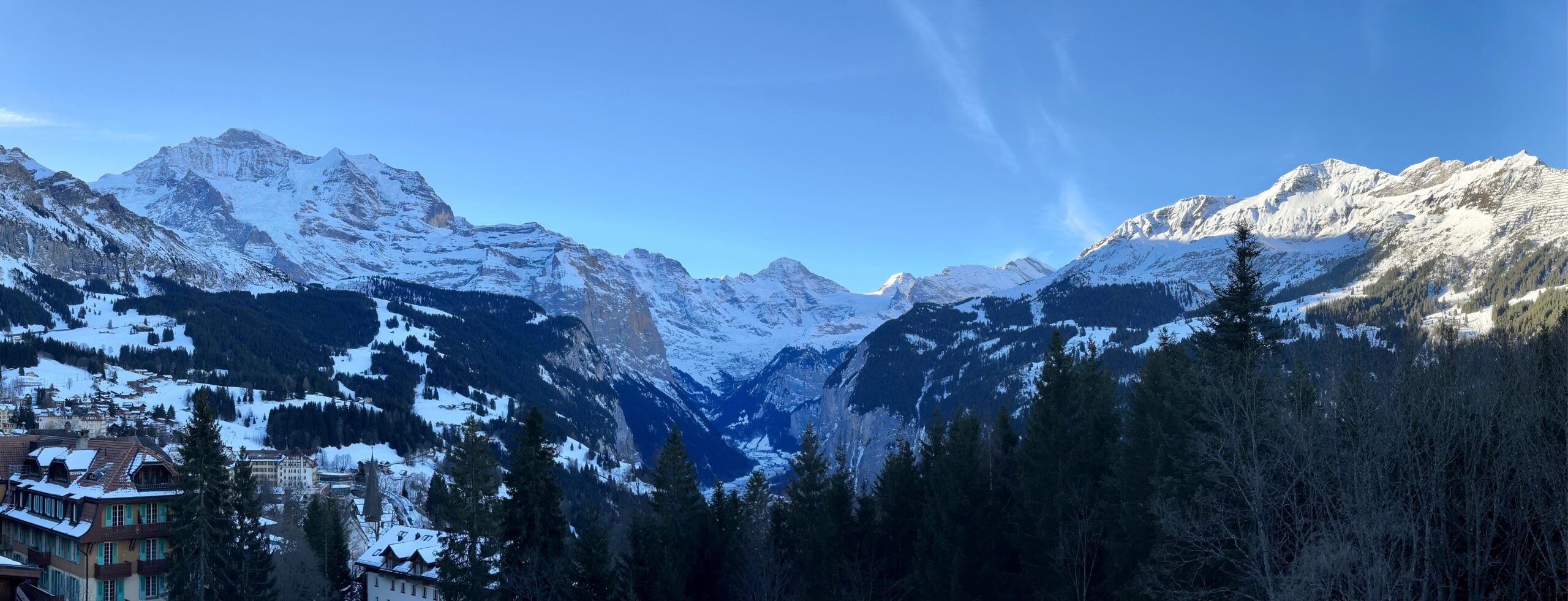 Fir trees, snow-covered buildings, snow-capped peaks under a blue sky.