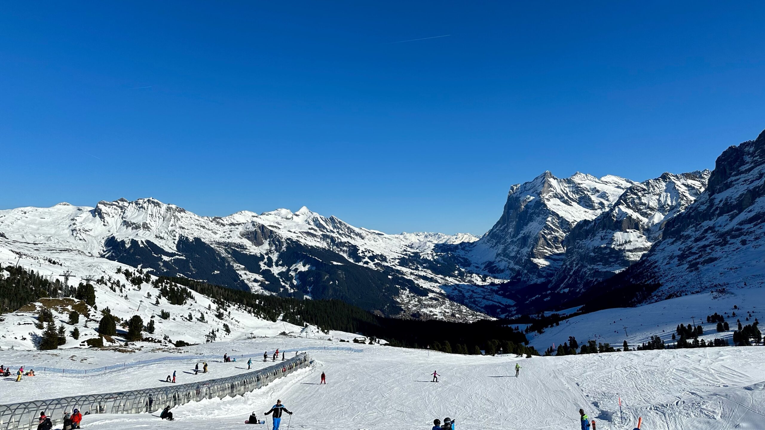 A wide, groomed ski piste against a backdrop of snow-covered mountains under a clear blue sky.
