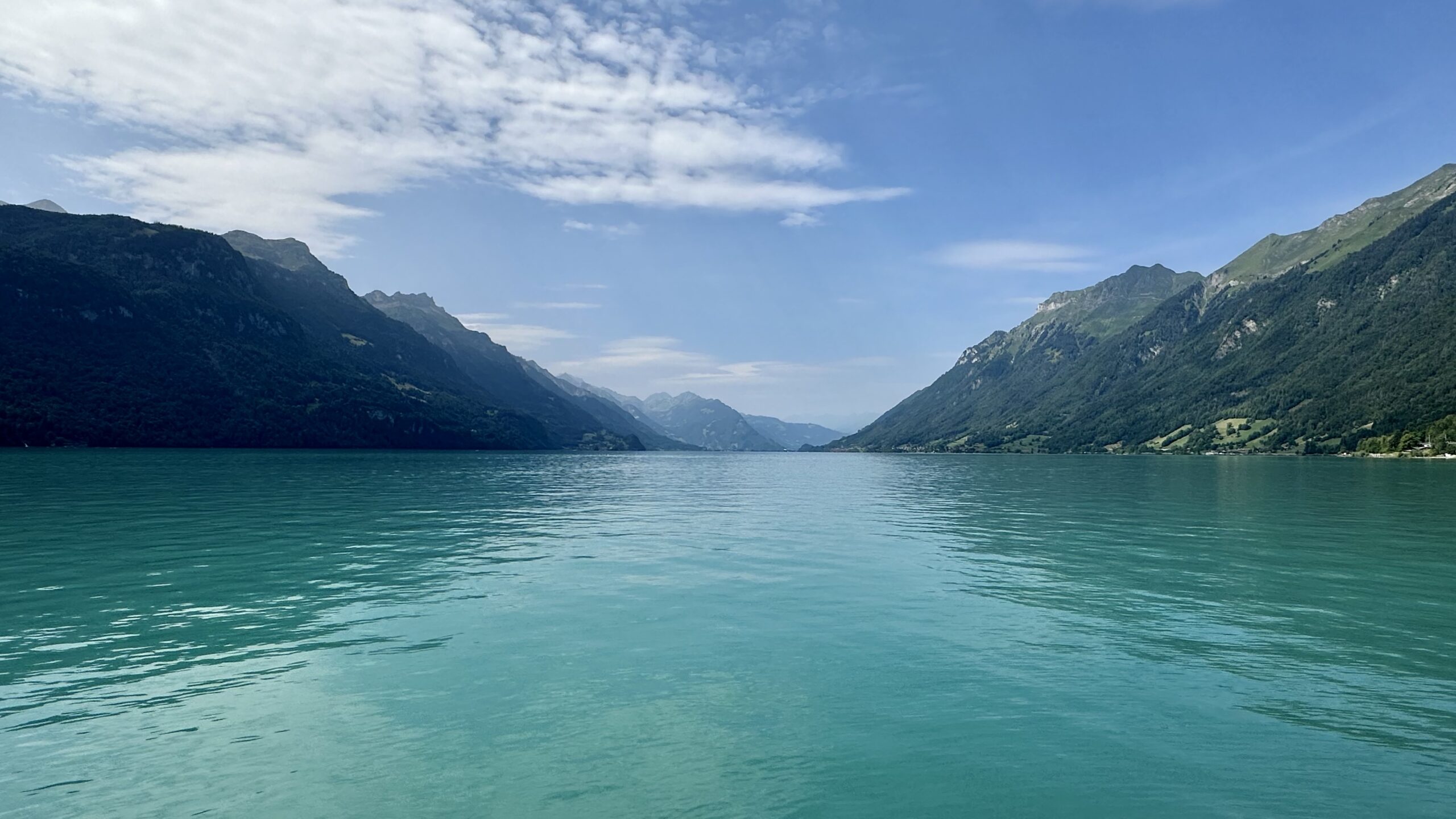 View of Lake Brienz, Switzerland. The water is greenish blue, forested mountains are in the distance and a blue sky with white clouds overhead.