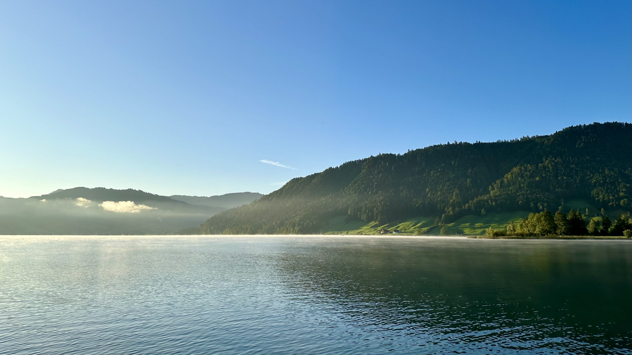 Green forested hills over a blue lake with a sheen of white mist under a blue sky.