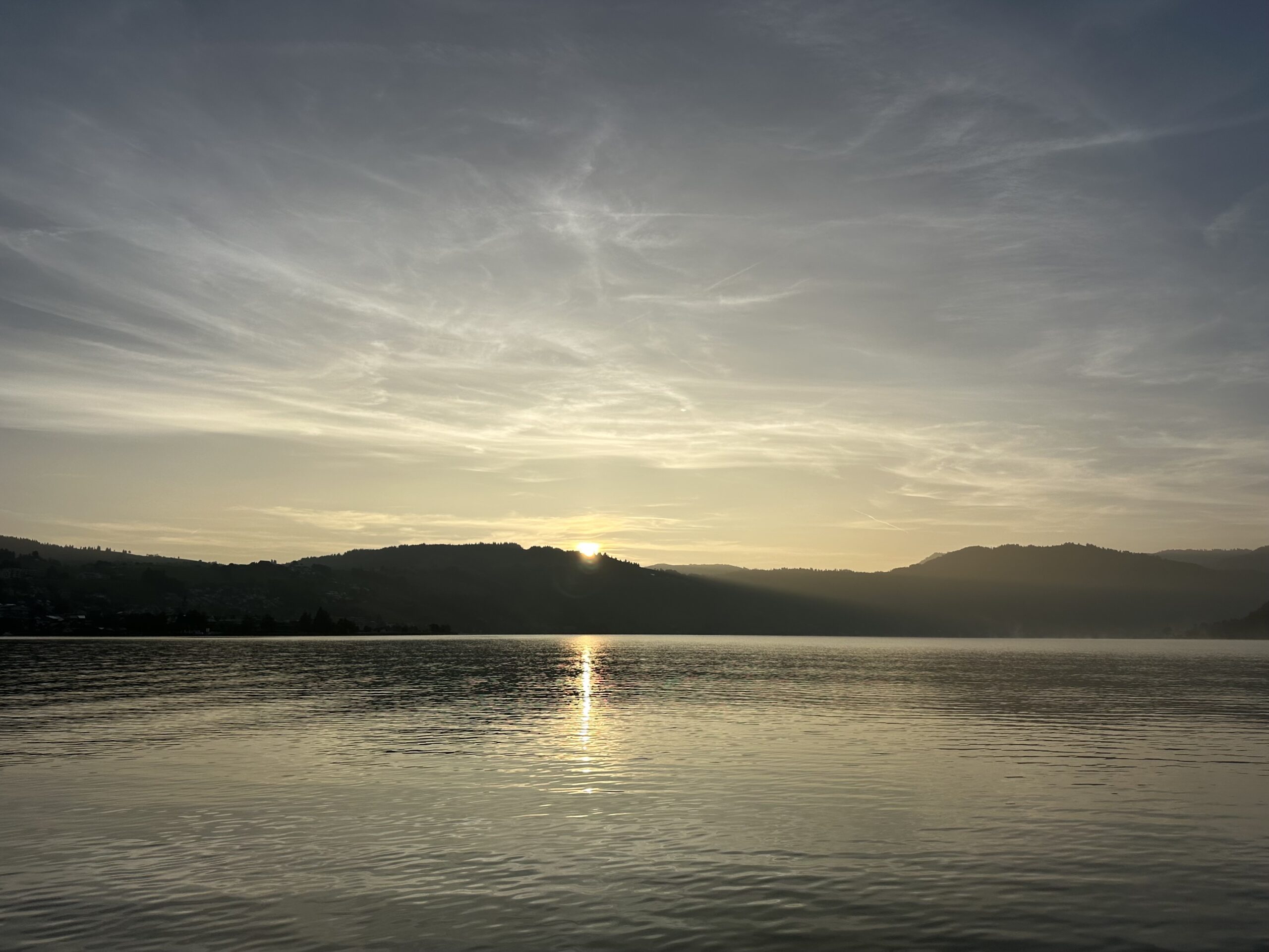 View across a lake directly into the sun rising over forested hills.