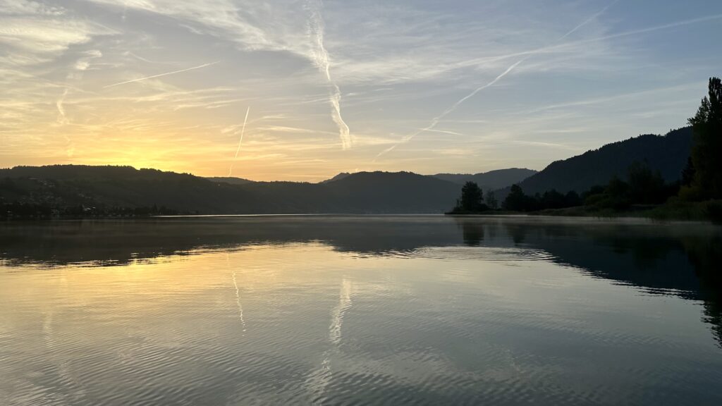 View of lake and hills taken from a SUP just before sunrise with a golden glow where the sun will soon appear. The lake has a few ripples but otherwise reflects the sky, which has more contrails than natural clouds.