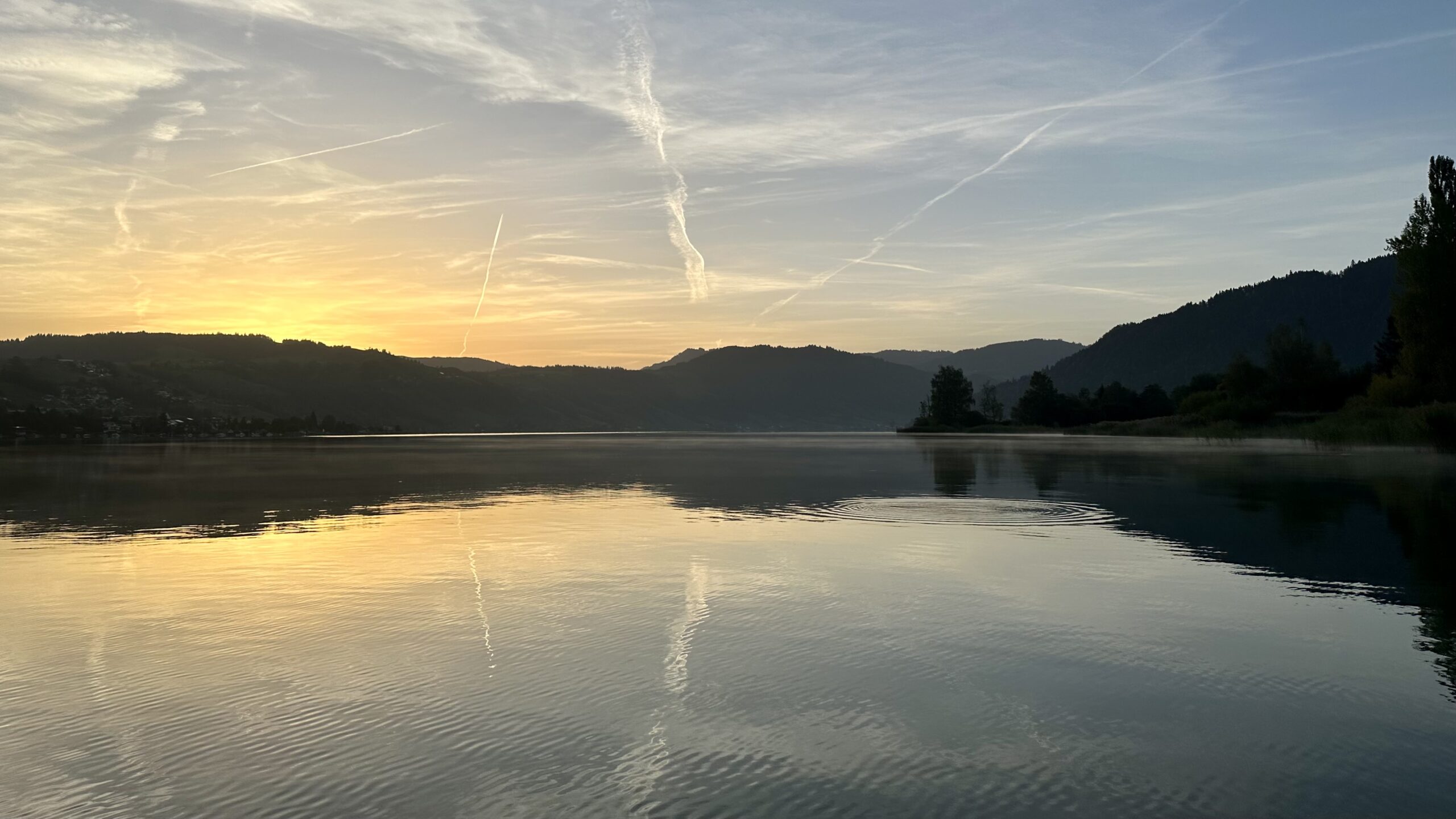 View of lake and hills taken from a SUP just before sunrise with a golden glow where the sun will soon appear. The lake has a few ripples but otherwise reflects the sky, which has more contrails than natural clouds.