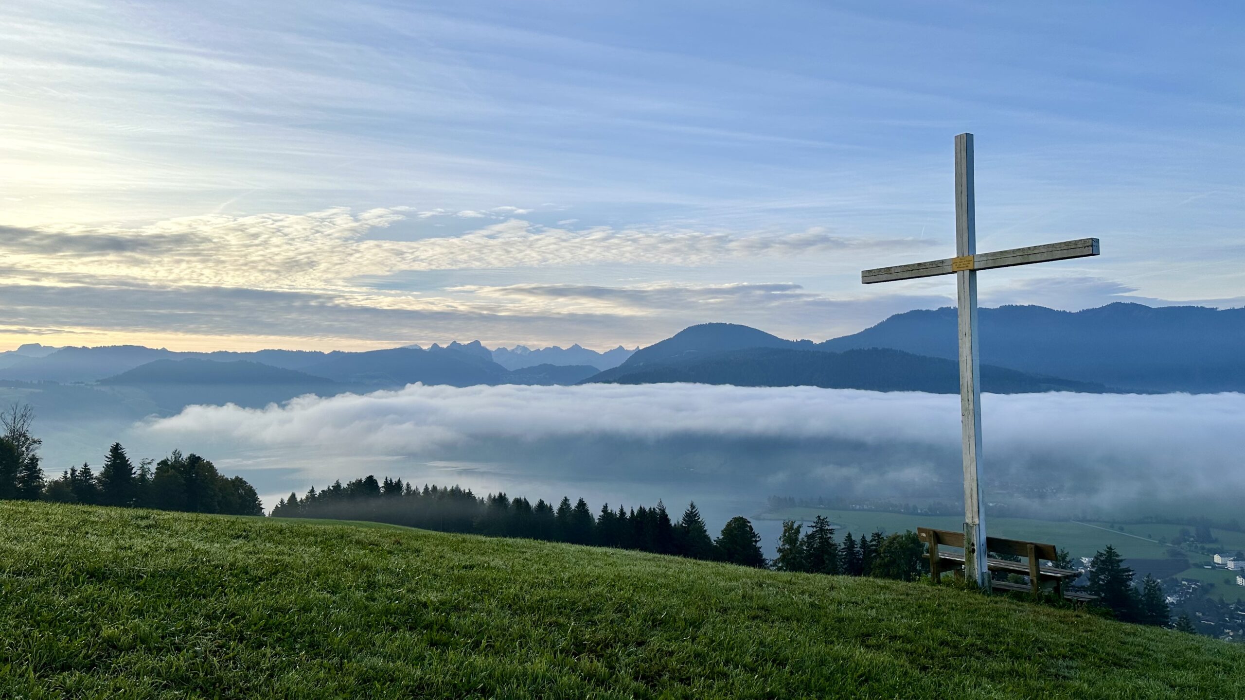 View of a cloud-filled valley with a lake. A large white cross loom over a park bench in the foreground on the right. Mountains line the horizon.