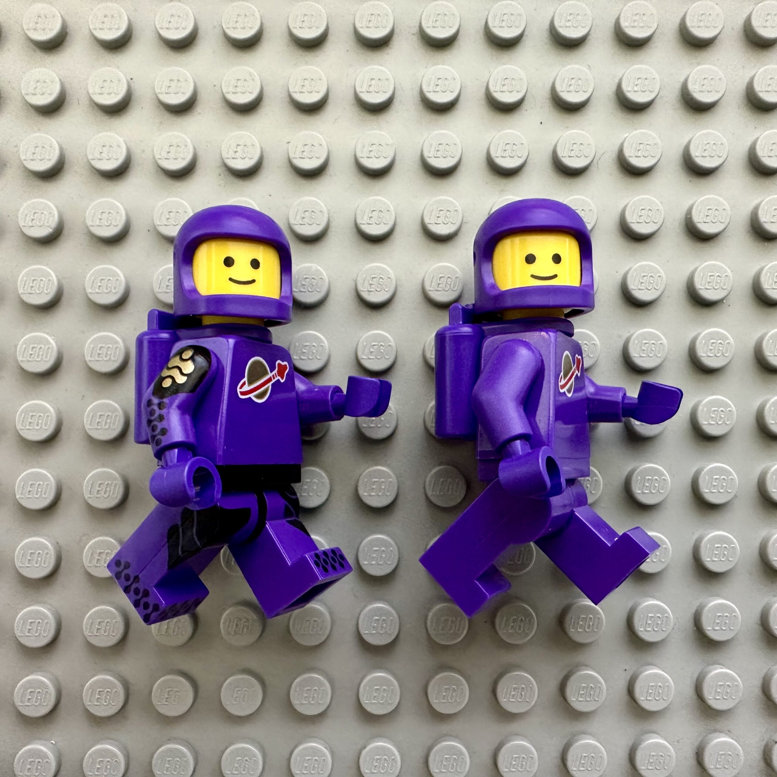 Two purple LEGO astronaut minifigs. The left figure has printed markings on its arms and legs while the right figure is plain.