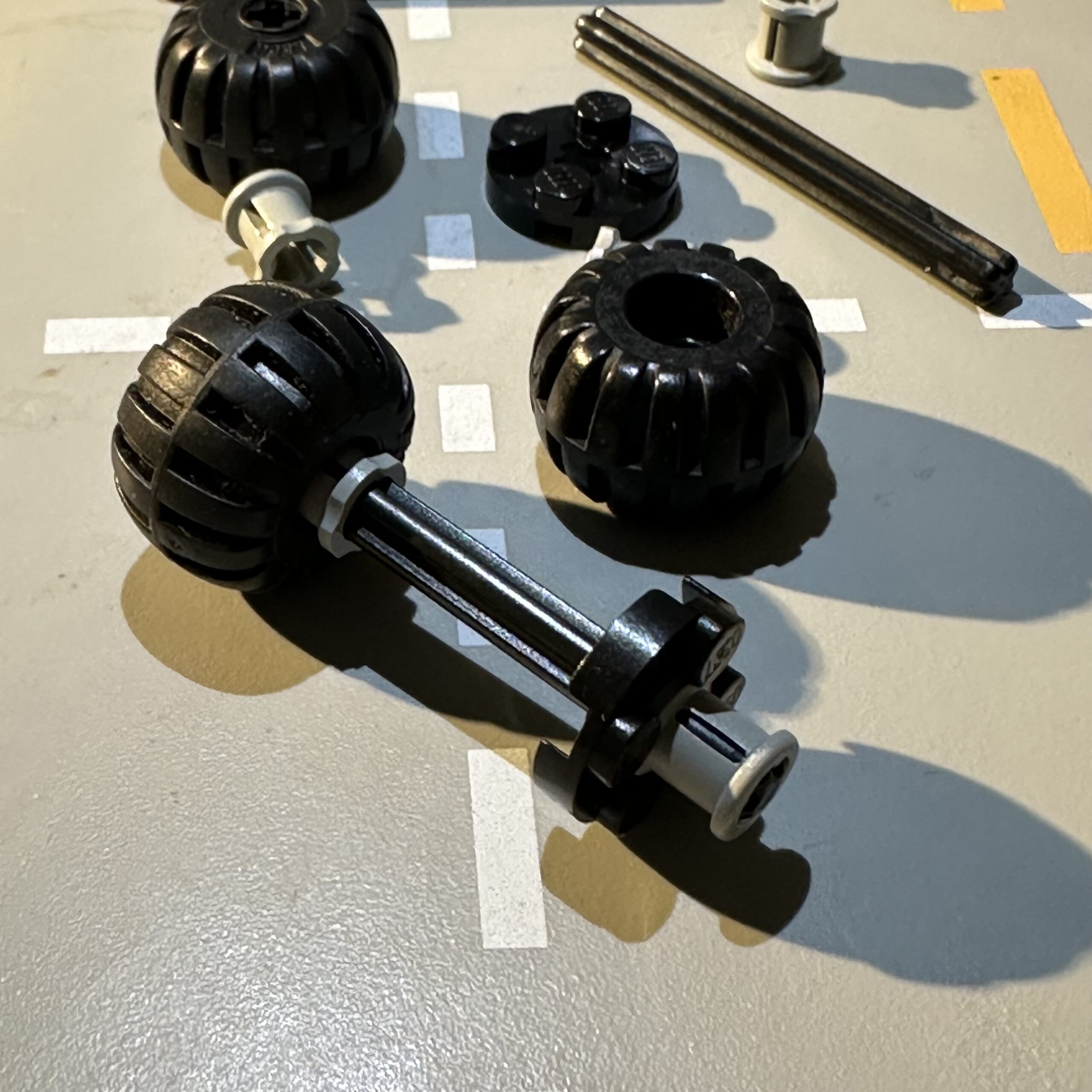 Collection of LEGO parts including a black rubber balloon tire piece connected to a black technic axle and secured with a gray bushing.