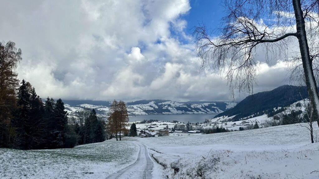 View of somewhat snowy valley with a gray lake. Some green is poking up from the grass. The sky is mostly cloudy with a small clear blue patch.