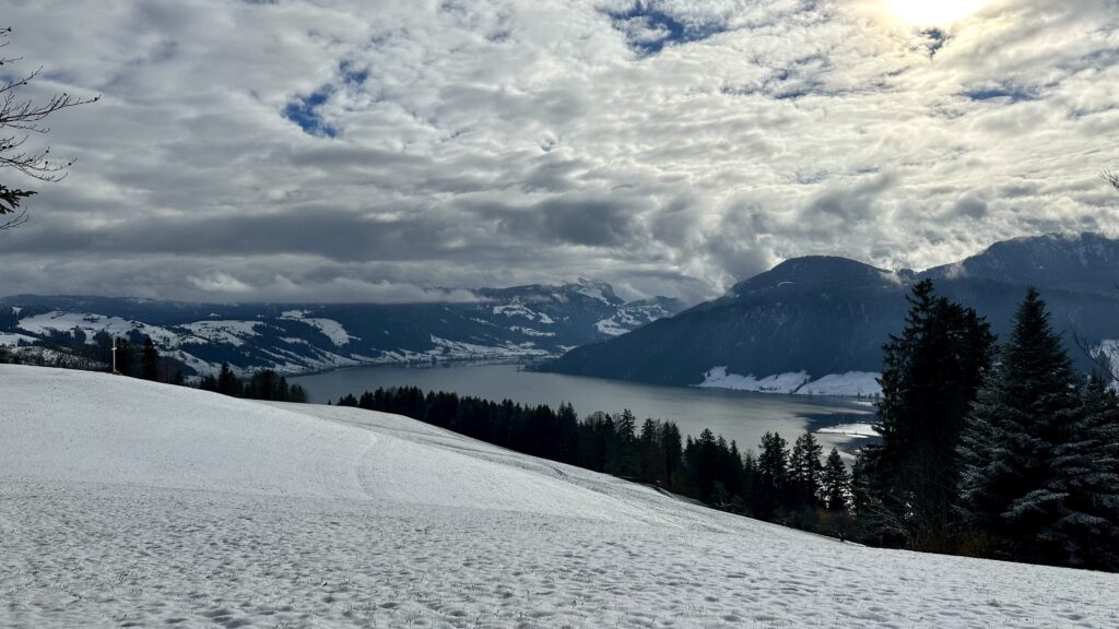 View from the Unterägeri White Cross on a day with snow on the ground. View of a lake surrounded by partially forested hills. The sky is filled with puffy clouds.