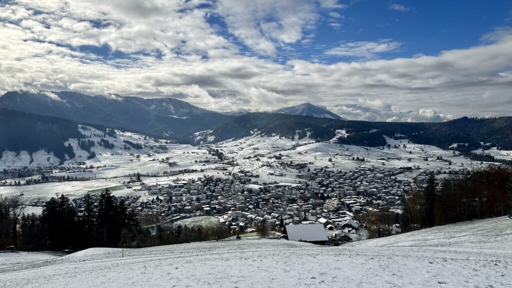 The town of Unterägeri in Canton Zug, Switzerland as seen from a hill on its northern side looking south. The valley is lightly covered in snow. The town is tightly clustered in the foreground with farmland in the distance giving way to forested hillsides that shape the valley. The sky is filled with puffy white clouds and a bit of blue.