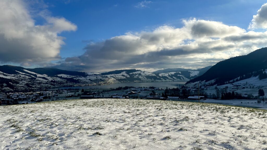 Partially snow-covered valley with a lake and a small town on its shores. The sky is blue with puffy clouds. Most of the valley is in shadow from the clouds.