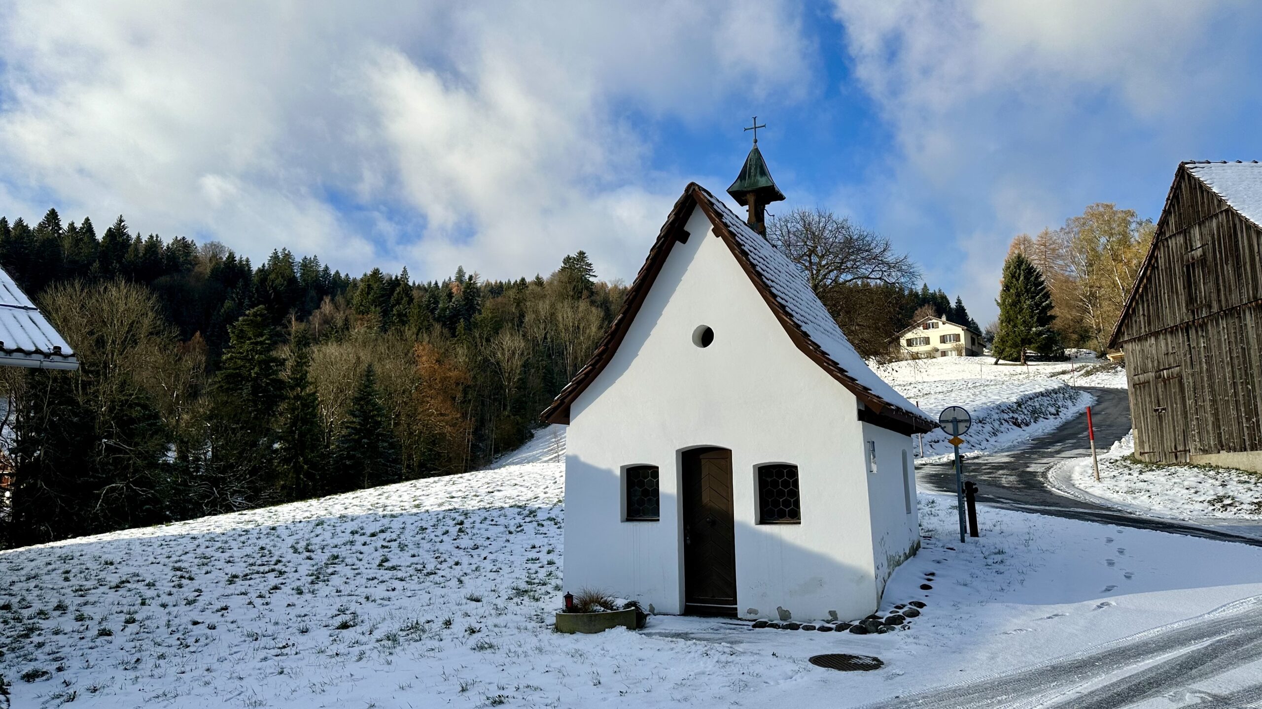 Small white chapel with a steep roof sitting on a snow-covered hillside next to a wooden barn.