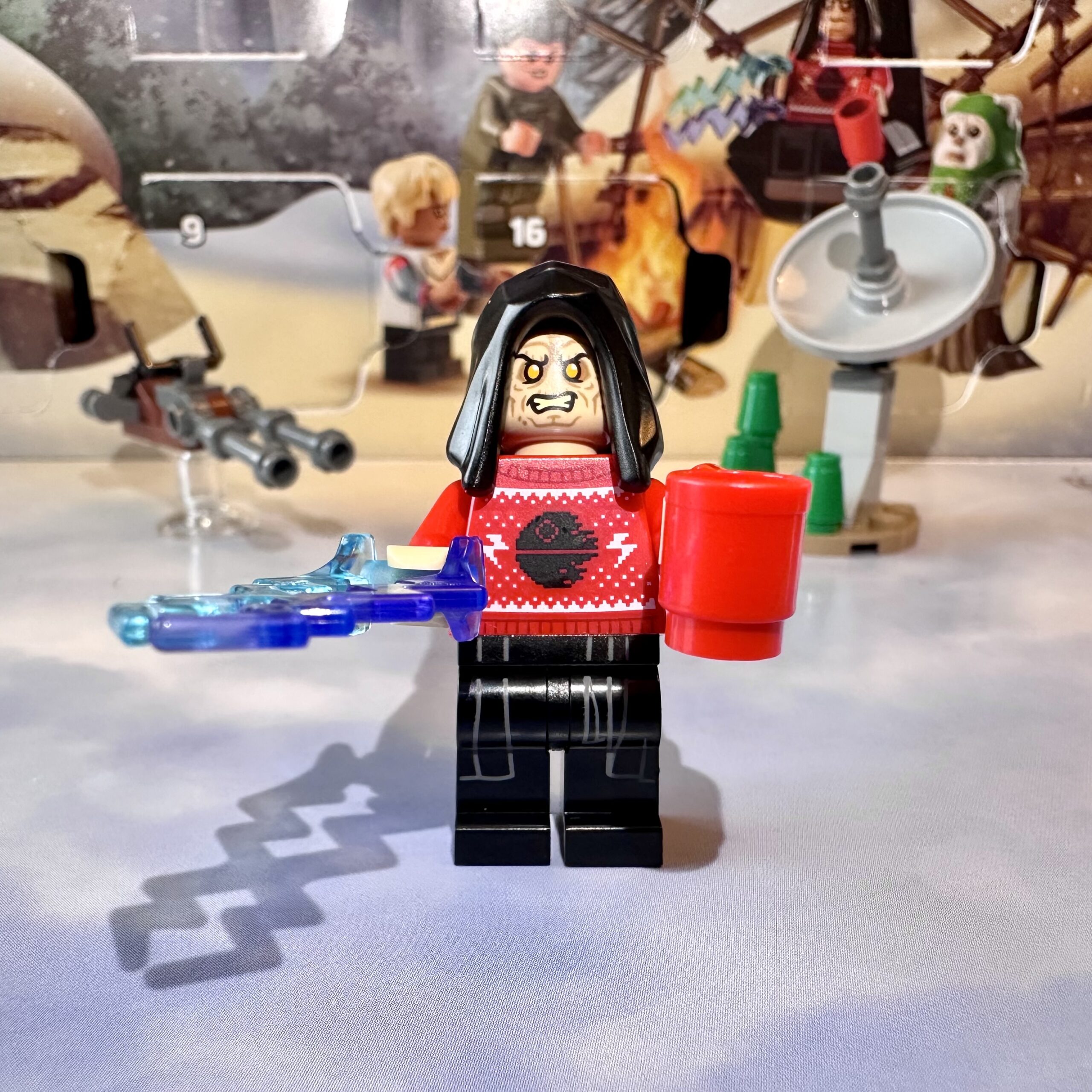 LEGO minifigure of Emperor Palpatine wearing an ugly red Christmas sweater with a Death Star under construction design on it. He holds a red coffee cup in his left hand and bolts of purple and blue energy fly from his right hand.