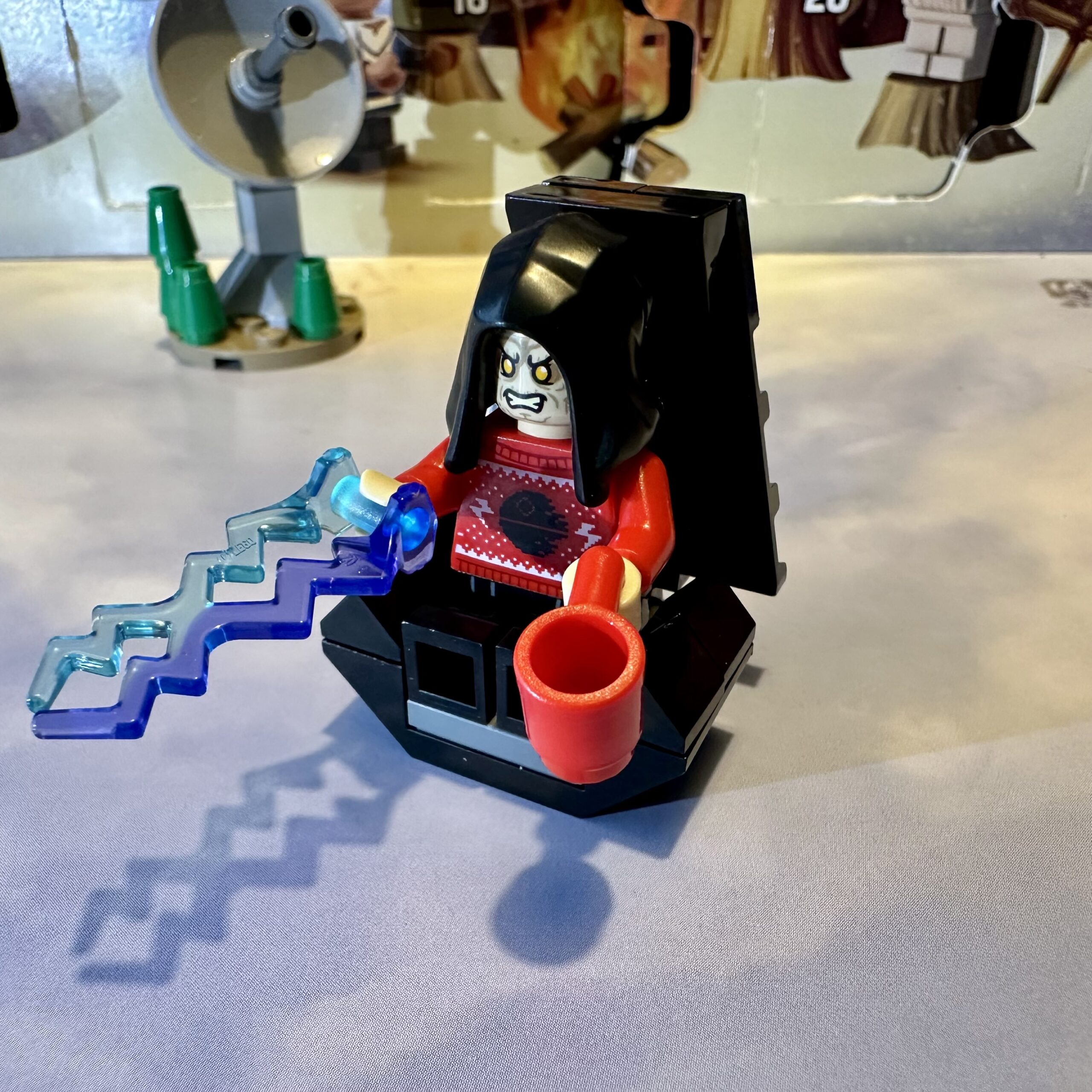 LEGO ugly Christmas sweater Emperor Palpatine minifigure seated in his high-backed black Imperial throne with a red coffee mug in his left hand and blue and purple energy bolts zapping out of his right hand.