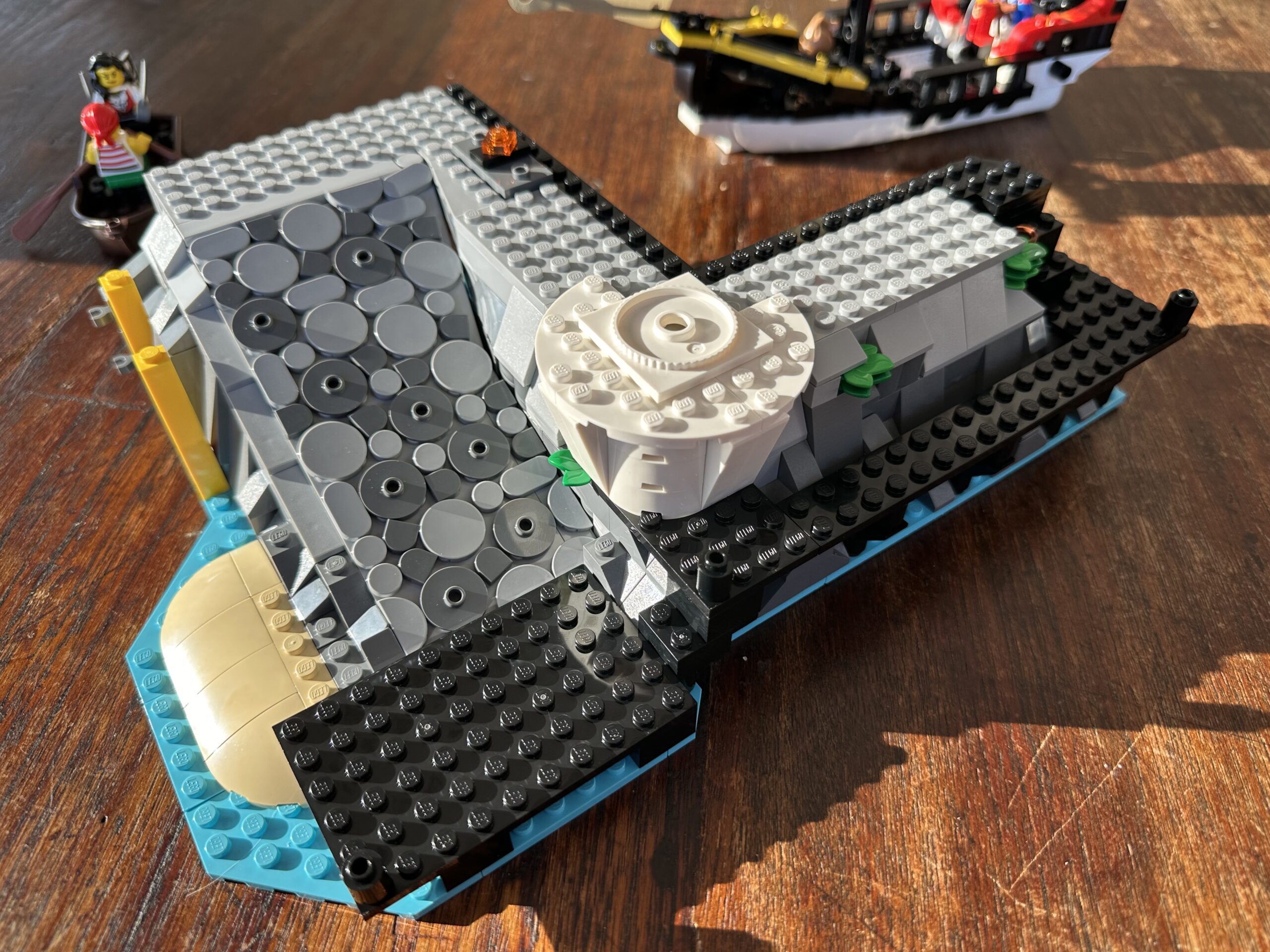 LEGO build in progress. A light and dark gray cobbled ramp leads from a black jetty. A bit of sand beach reaches into turquoise waters to the left.
