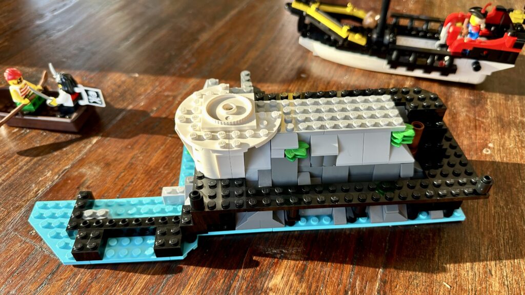 LEGO build in progress. Turquoise waters, black jetty, darkand light gray craggy rocks, and some white brick foundations. A few green leaves peek out amongst the rocks.