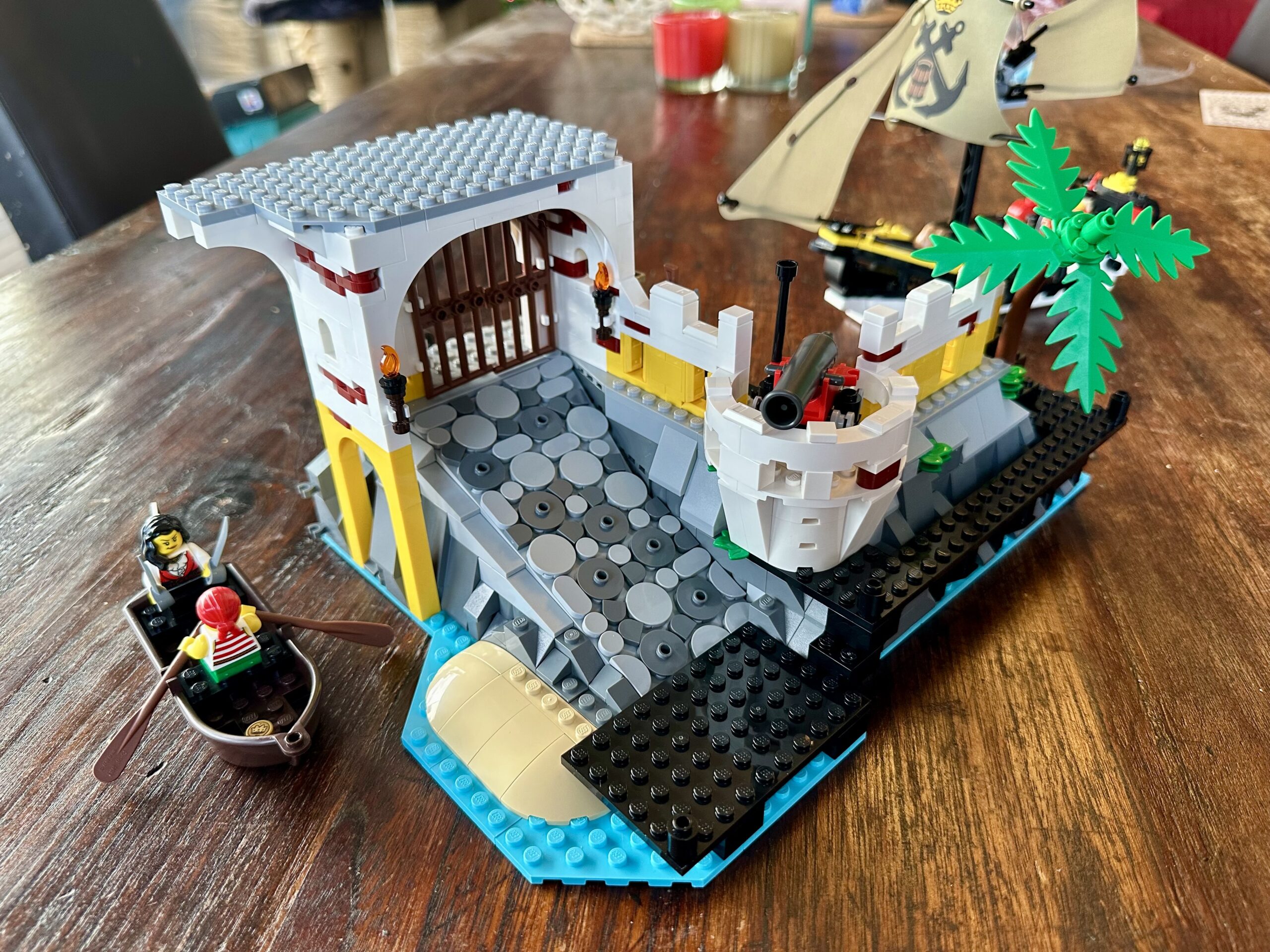 LEGO build of set 10320 Eldorado Fortress in progress. The first floor of a gatehouse sits at the top of a ramp that leads up from the jetty. There's a palm tree on the right and a rowboat crewed by two pirates on the left.