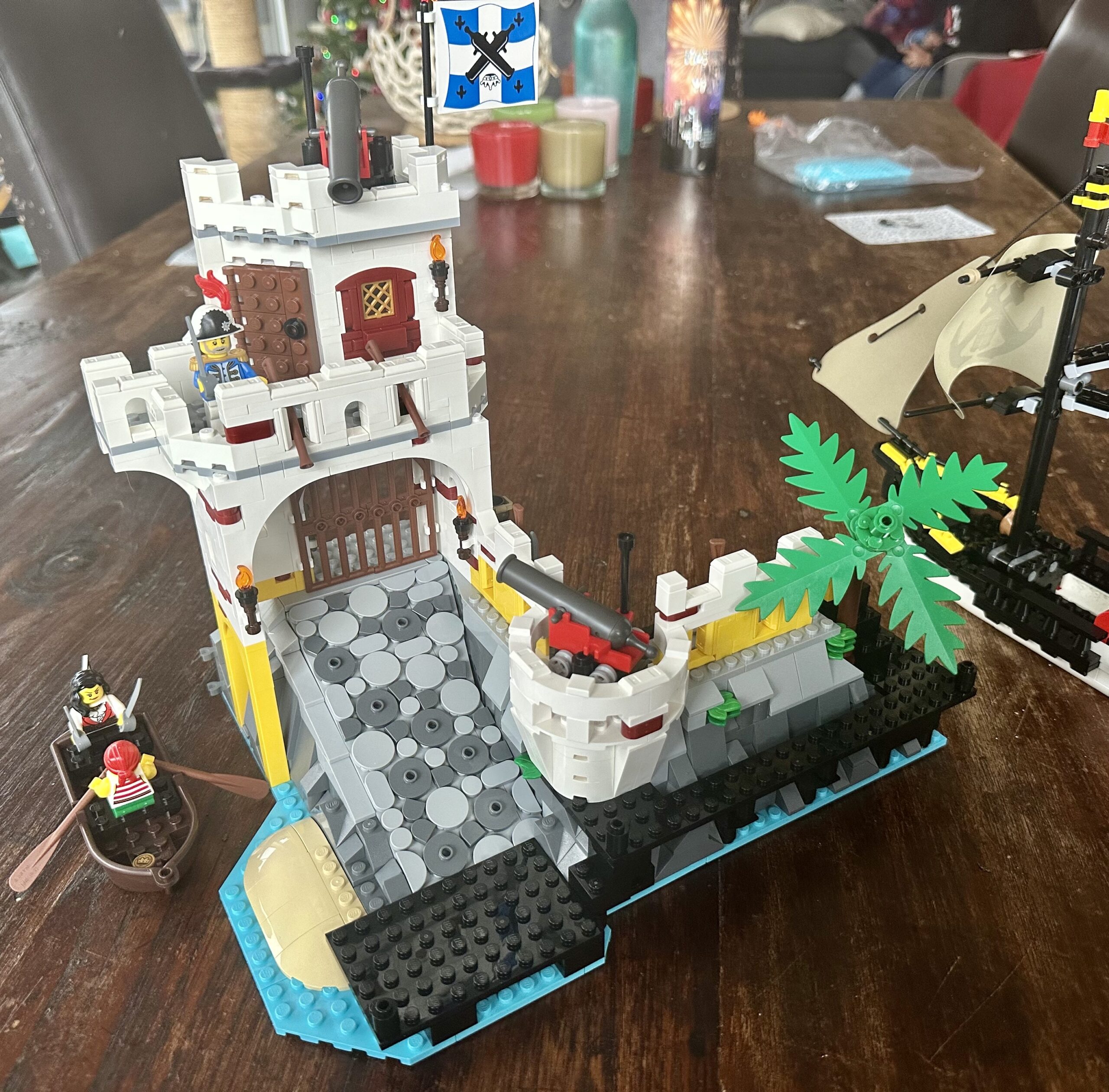 LEGO build of set 10320 Eldorado Fortress in progress. A second story has been added to the gatehouse. It features a balcony with two rifles pointed down at the ramp leading to the gate, a small office with a cannon mounted above it, and a blue and white flag with crossed cannons. An officer stands on the balcony brandishing a cutlass.