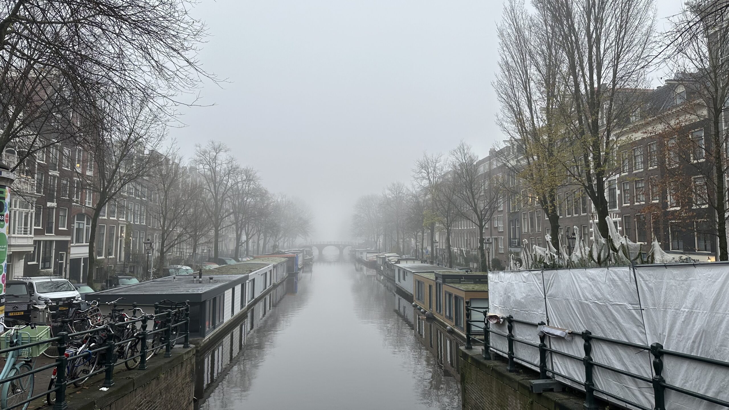 View down an Amsterdam canal on a foggy morning. The canal is flanked by house boats, leafless trees, and narrow row houses.