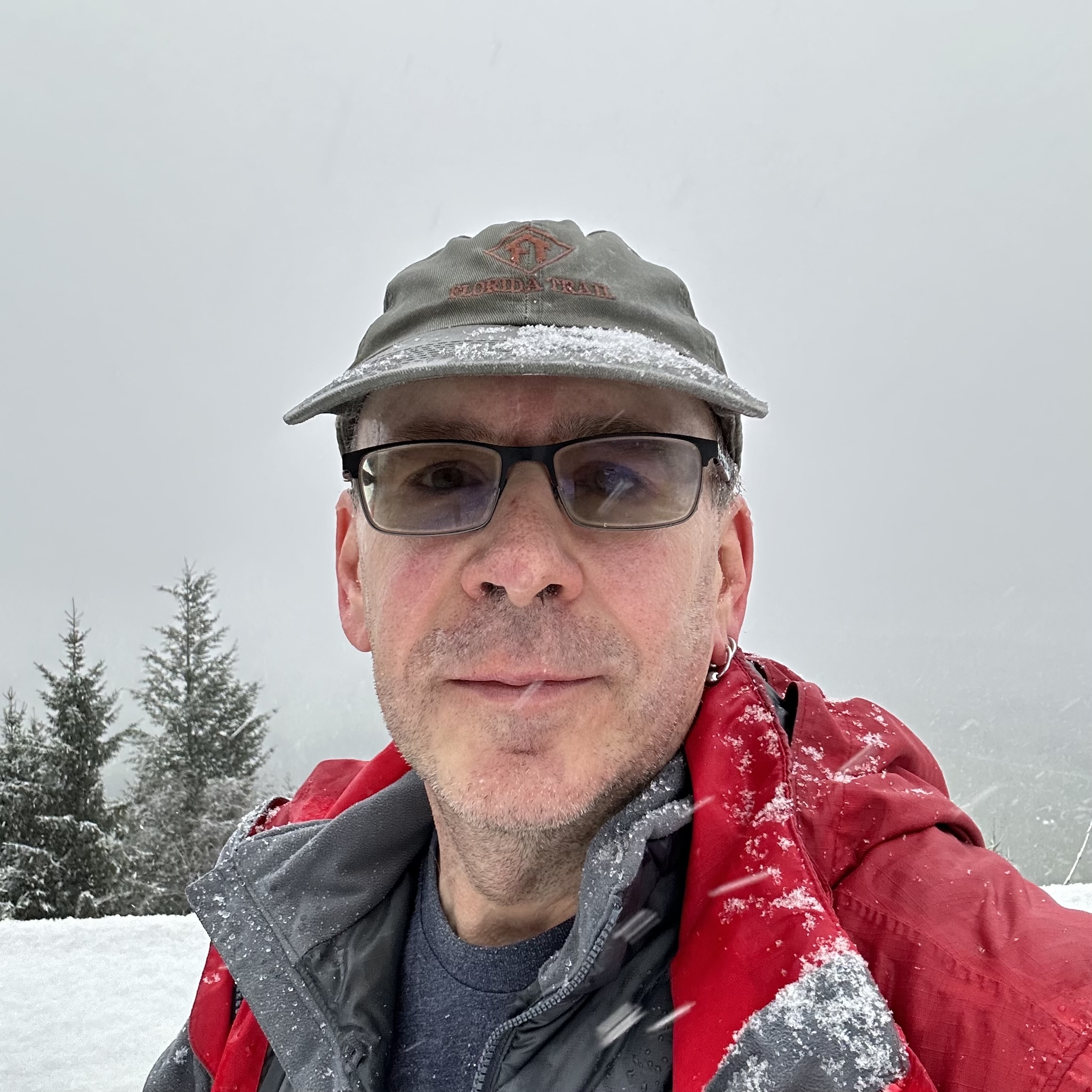 Selfie of middle-aged white guy in glasses, red ski jacket and an olive Florida Trail baseball cap. He's a bit covered in snow.