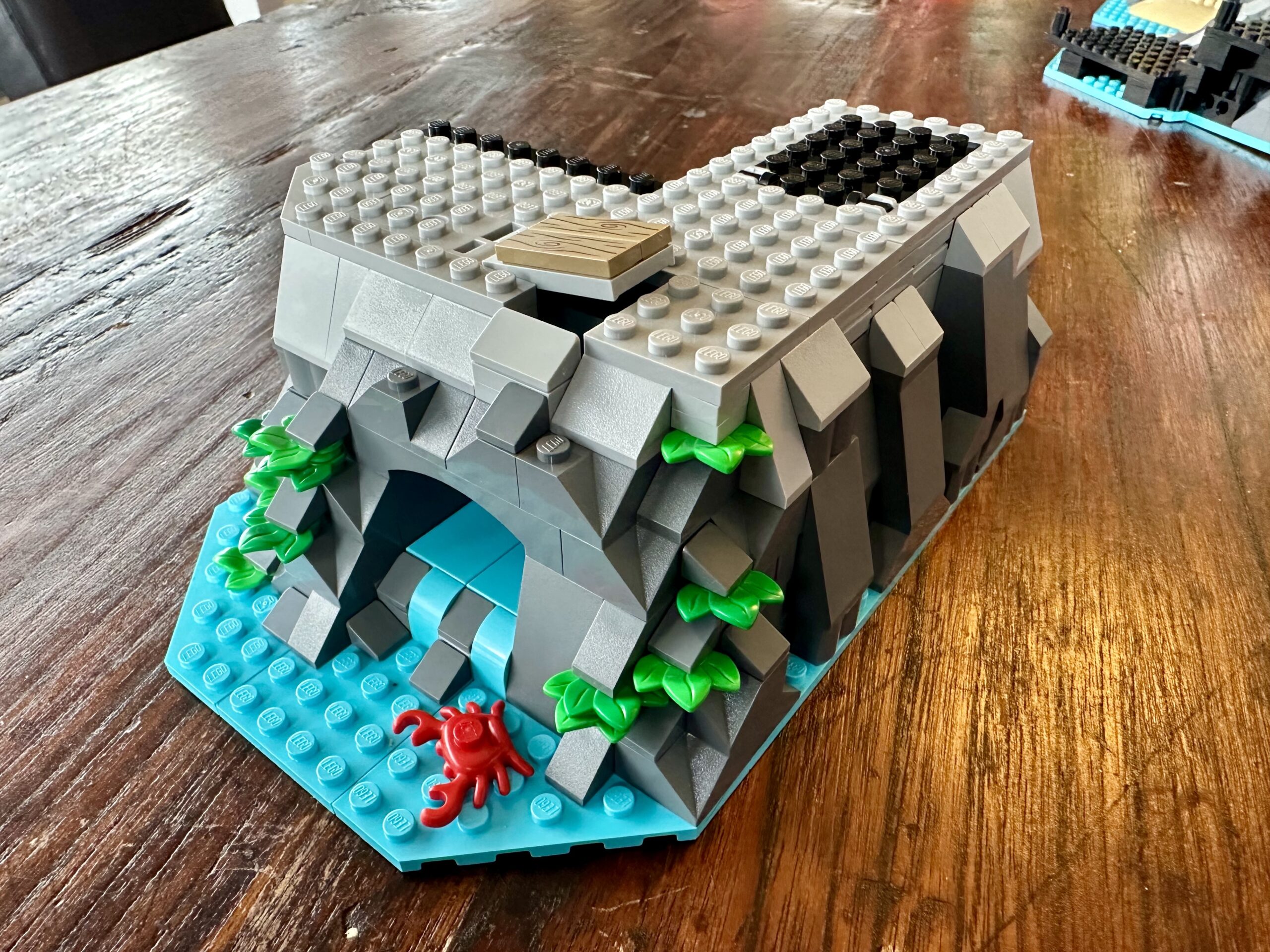 LEGO build of set 10320 Eldorado Fortress in progress. An L-shaped section of dark and light gray cliffs speckled with green foliage. Some water flows out from a cave to the turquoise waters of the beach. A red crab is in the water.