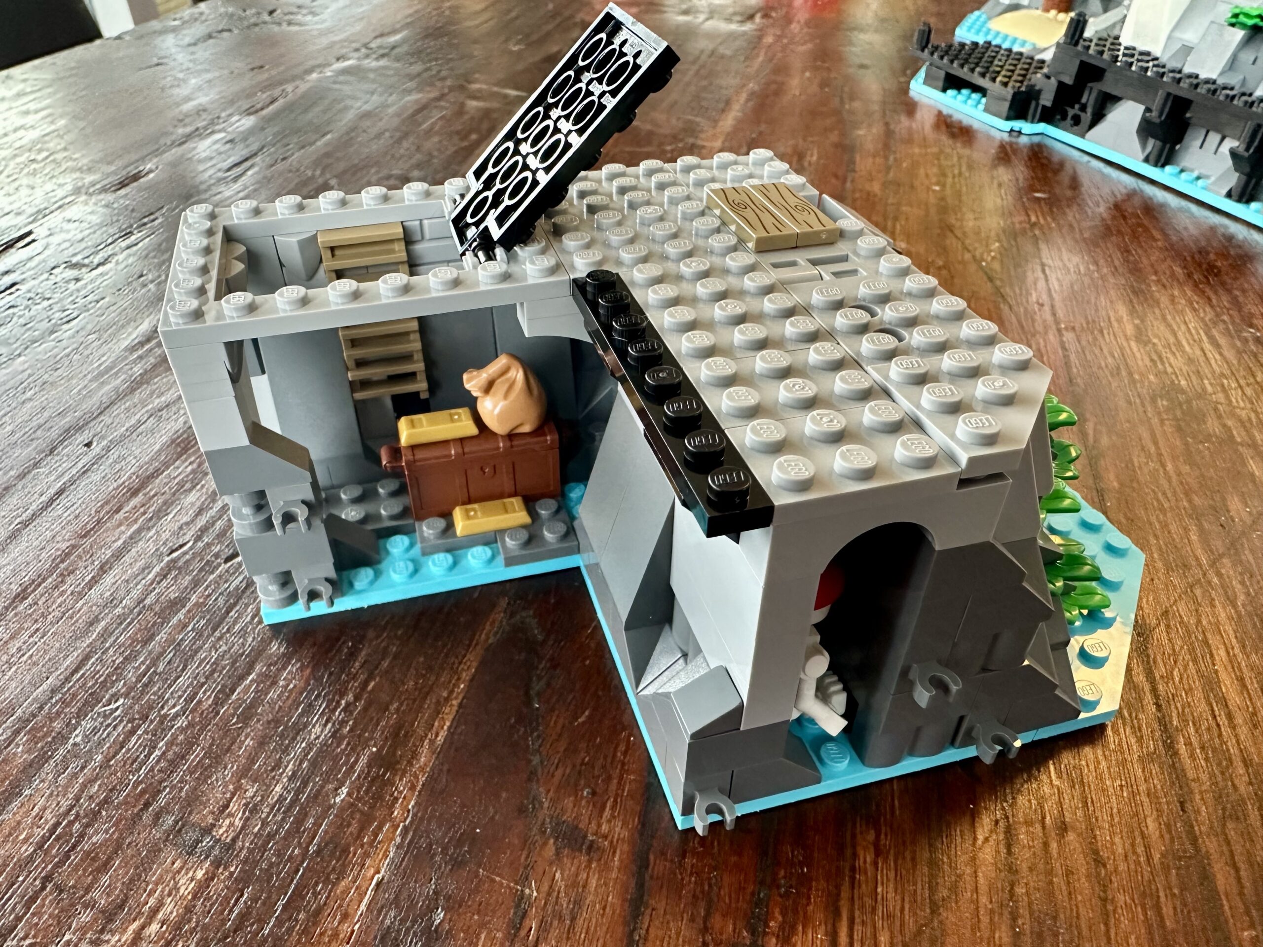 LEGO build of set 10320 Eldorado Fortress in progress. An L-shaped section of gray stone foundations. There's a treasure room with gold ingots accessed by ladder and trap door. To the right a skeleton wearing a red bandana is partially visible, wedged into a narrow crevice in the rocks.