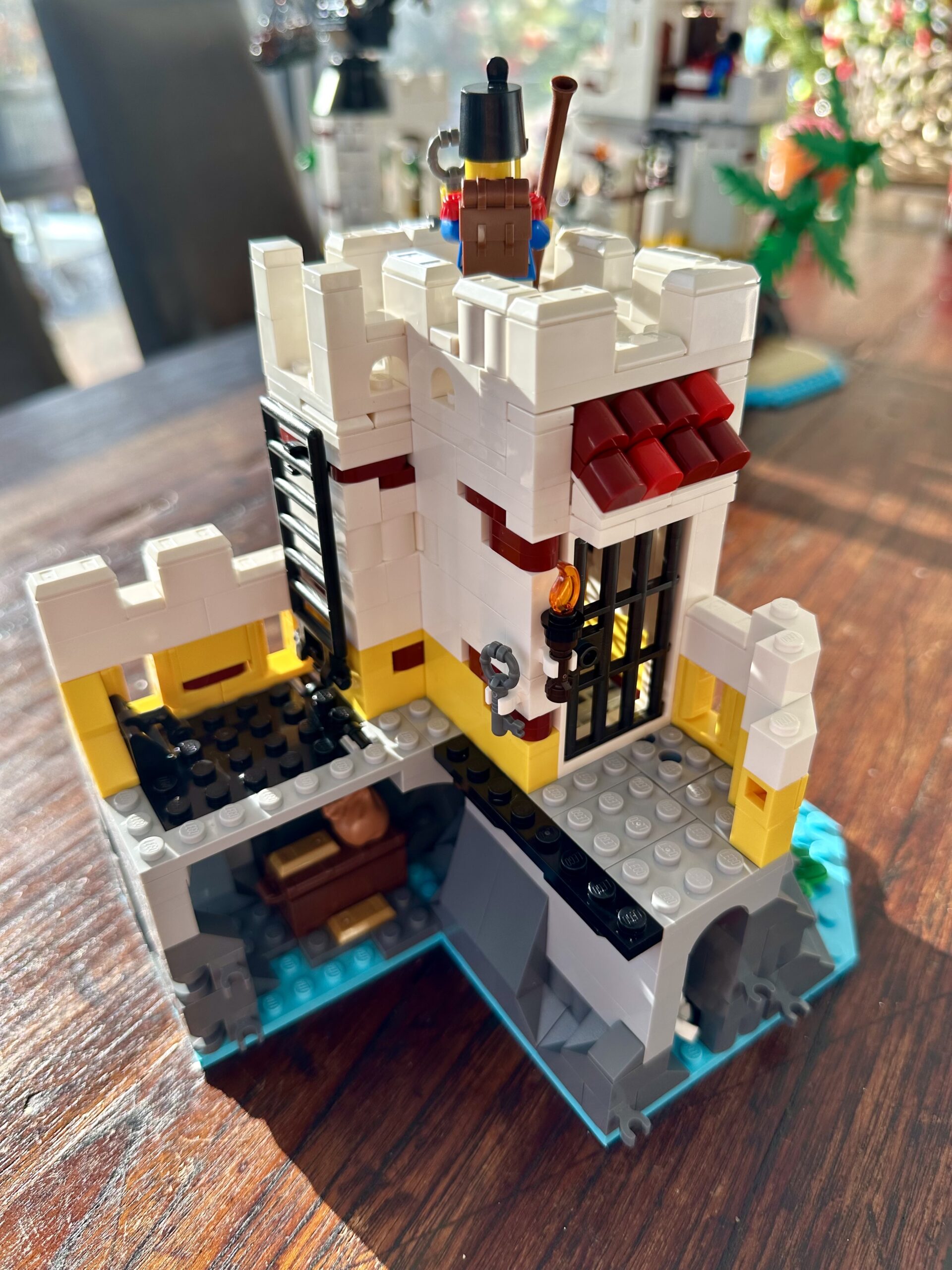 LEGO build of set 10320 Eldorado Fortress in progress. An L-shaped section with gray rock foundations and a white tower with yellow and dark red accents built atop it. The tower holds a prison cell and a soldier stands guard up top (facing away).