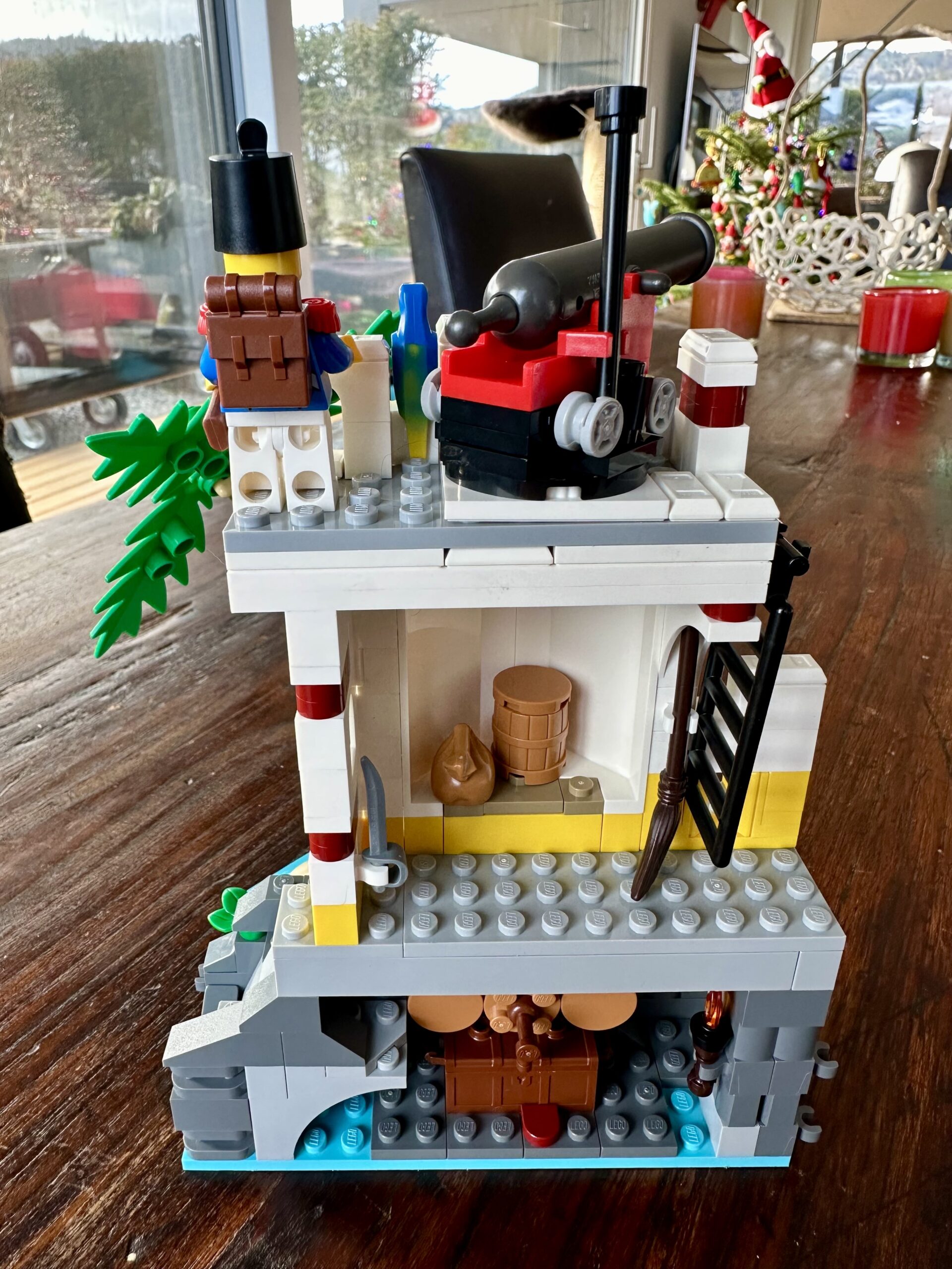 LEGO build of set 10320 Eldorado Fortress in progress. The rear side of a white tower revealing a seaside grotto with treasure chest and three kegs, a store room with weapons, barrel, and sack, and a roof with a cannon and an Imperial guard.