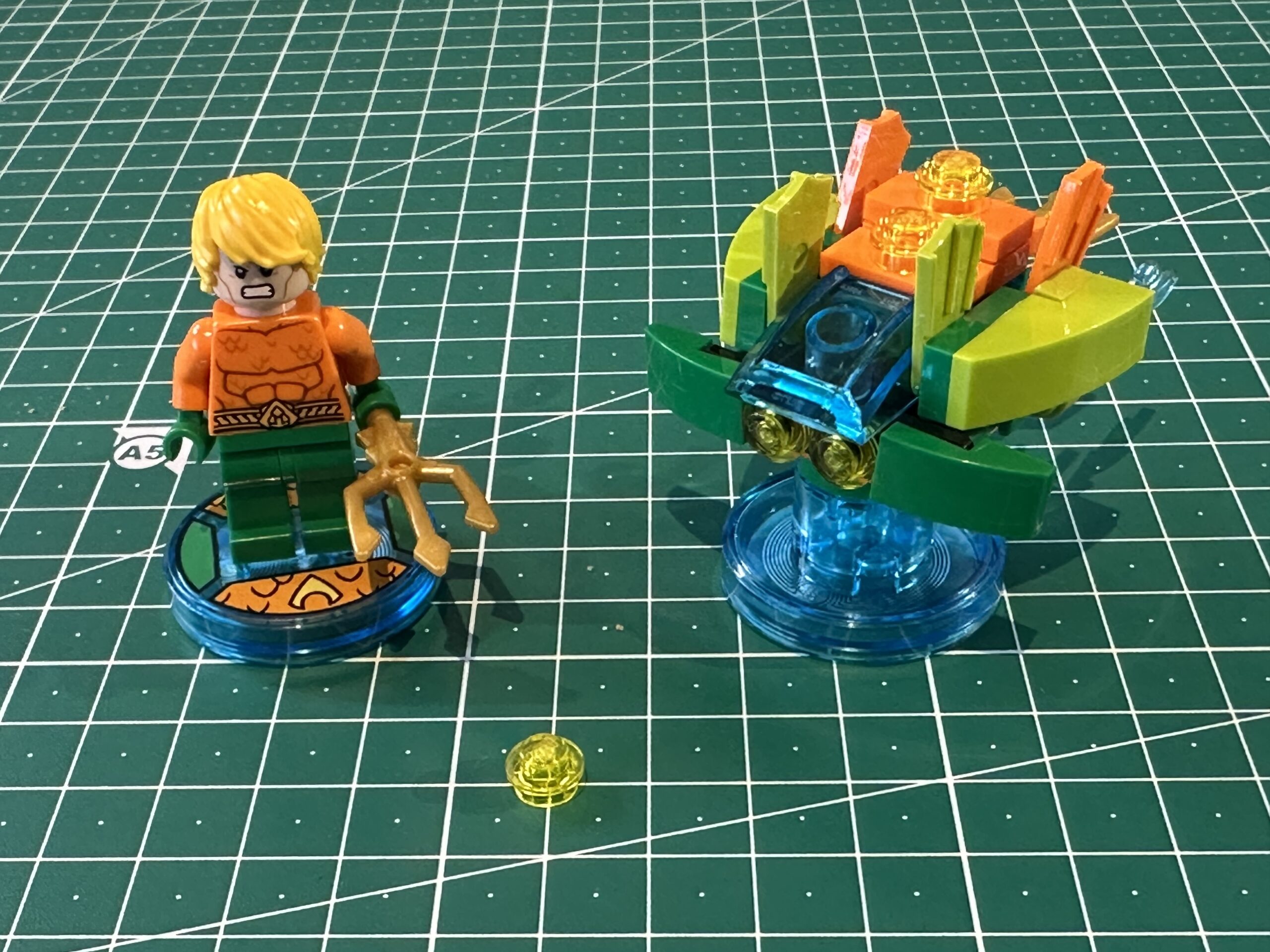 LEGO minifigure of Aquaman in his signature orange and green outfit holding a golden trident on the left and some weird orange and green underwater scooter on the right.