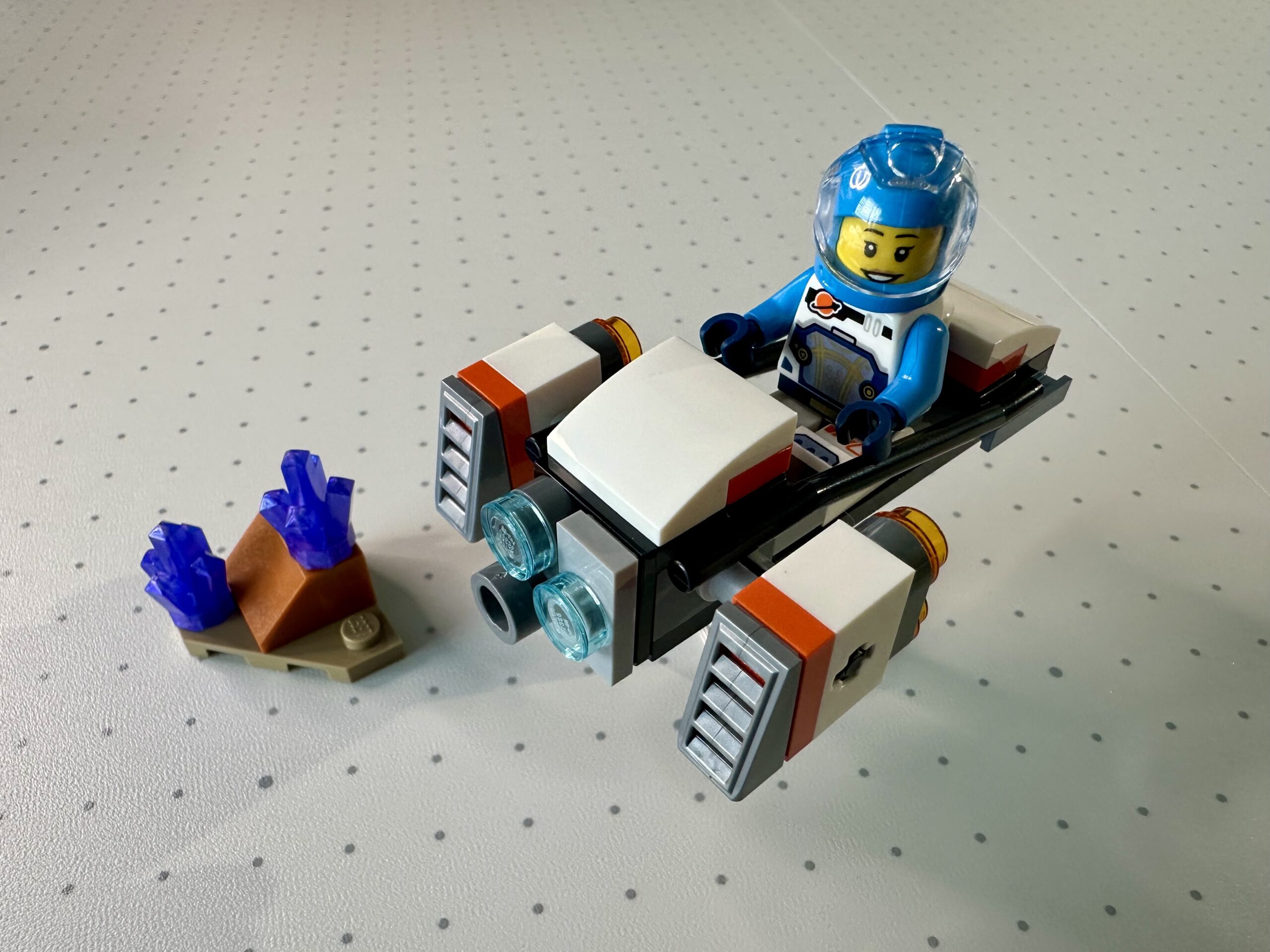 LEGO hoverbike predominantly in white, dark gray, and reddish orange. Piloted by an astonaut in an azure and white spacesuit. Nearby is a small corner of dark tan terrain sprouting a couple of purple crystals.