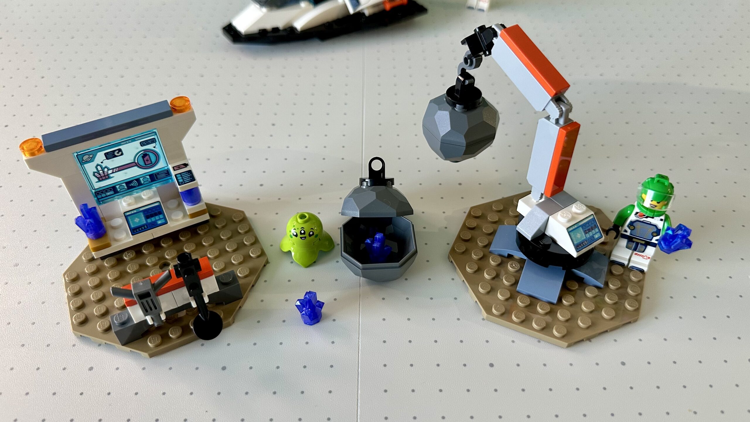 On the left is a science station with computer screen, a purple crystal, and a battery pack. In front of it is a small rack holding some tools. On the right is a crane lifting an asteroid. An astronaut in green and white stands next to it holding a purple crystal. In between are an asteroid cracked open to remove purple crystals inside and a chunky happy lime green alien.