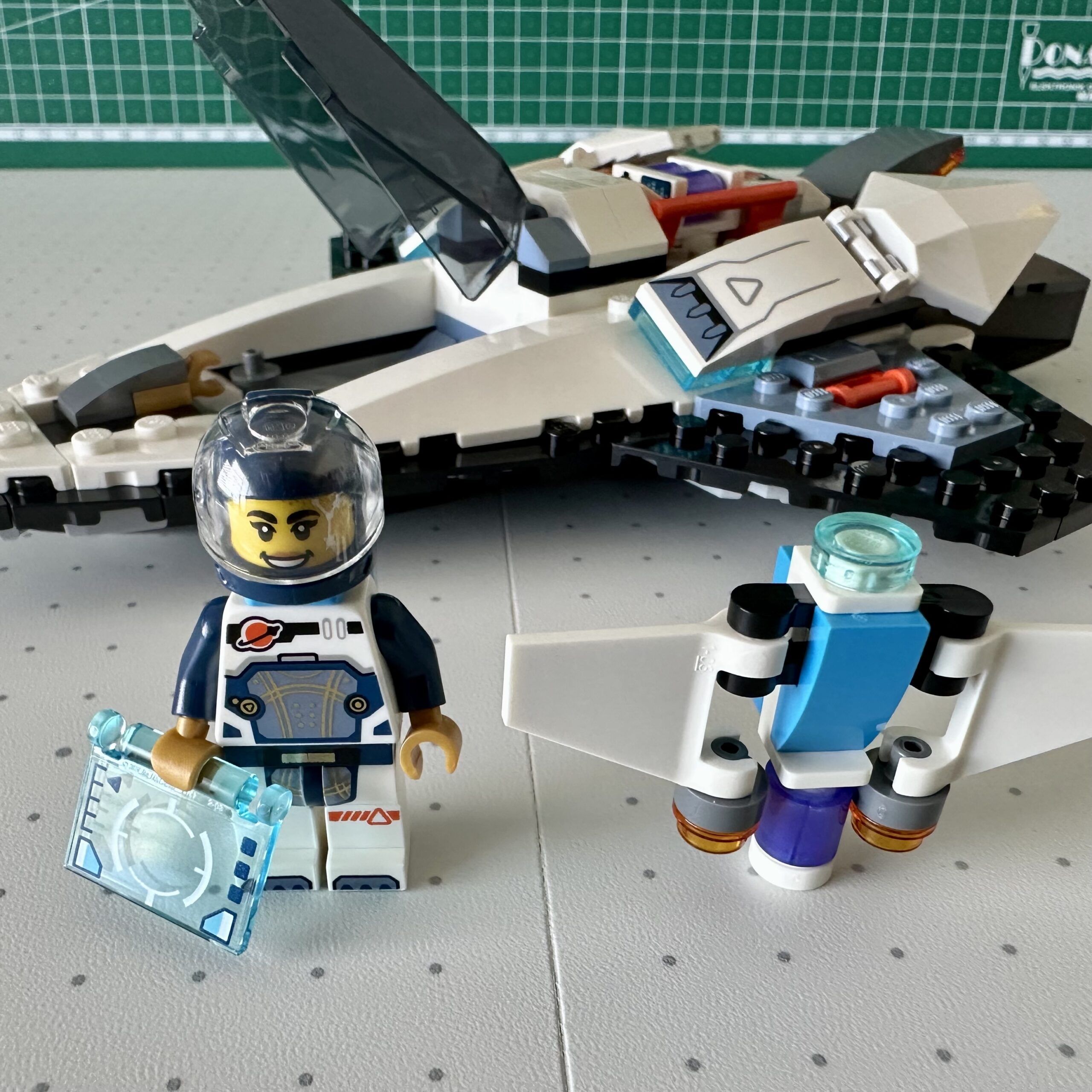 A female astronaut LEGO minifigure with dark blue helmet and sleeves holds a transparent blue panel with computer display. A brick-built jet pack /drone stands next to her. In the background her spaceship is parked with the canopy open.