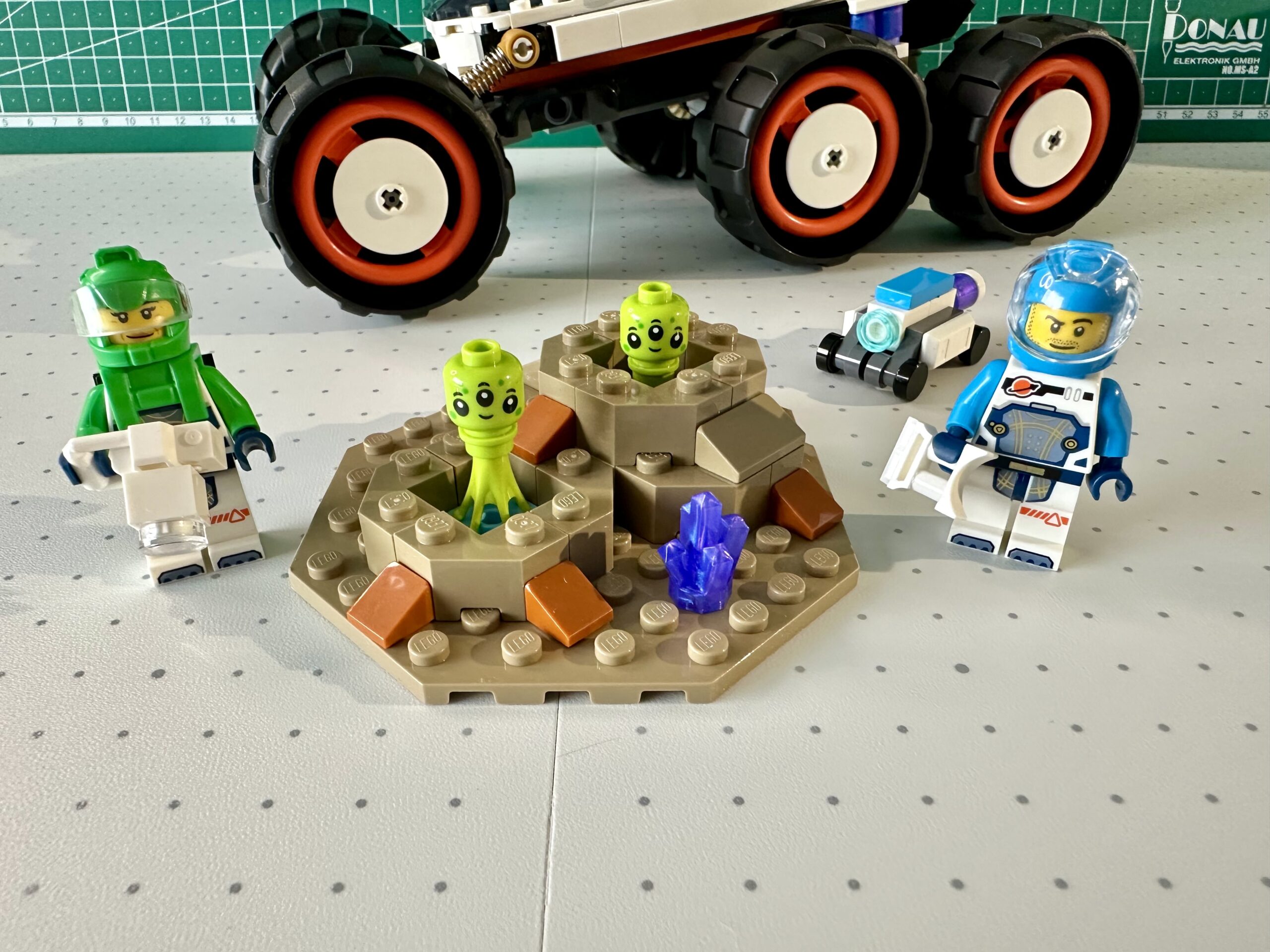 Green and azure-suited LEGO astronauts and a small wheeled robot flank a section of dark tan terrain with two craters. Happy looking 3-eyed lime green aliens sit in each crater.