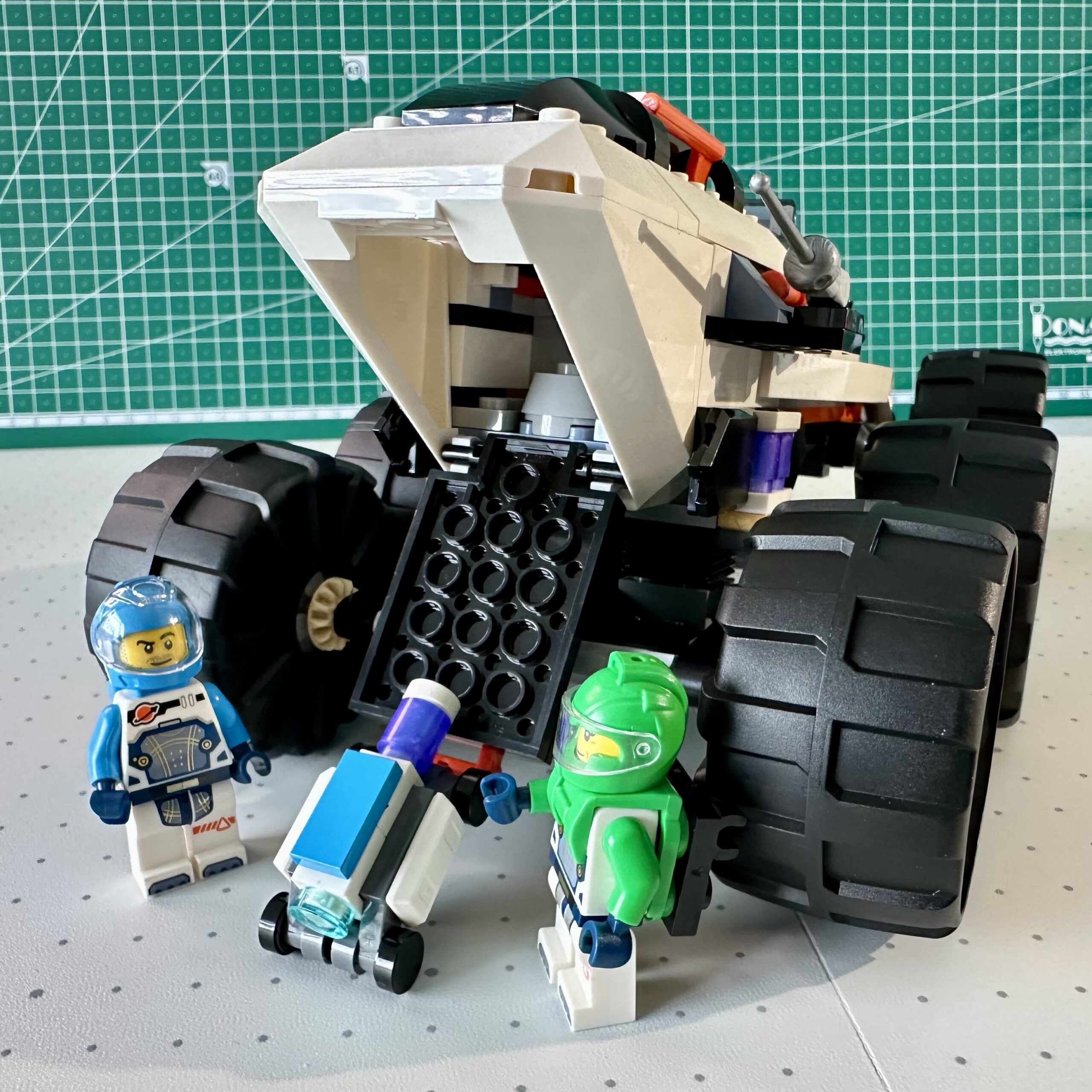 Two LEGO astronauts help a small wheeled robot move down the back ramp of their large monster truck space rover. Sadly for the little robot the ramp doesn't even reach the ground because the rover sits so high.