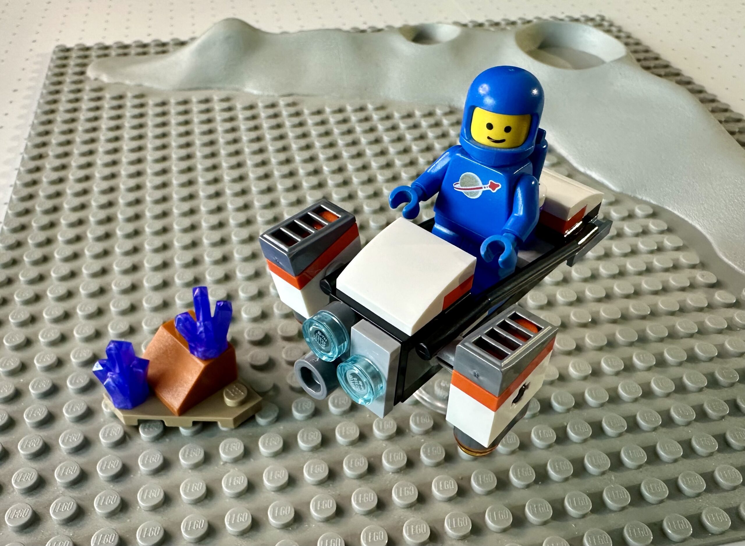 A classic blue LEGO spaceman riding the hover bike from the 2024 Space hover bike. Posed on a classic gray crater baseplate from the 1970's.