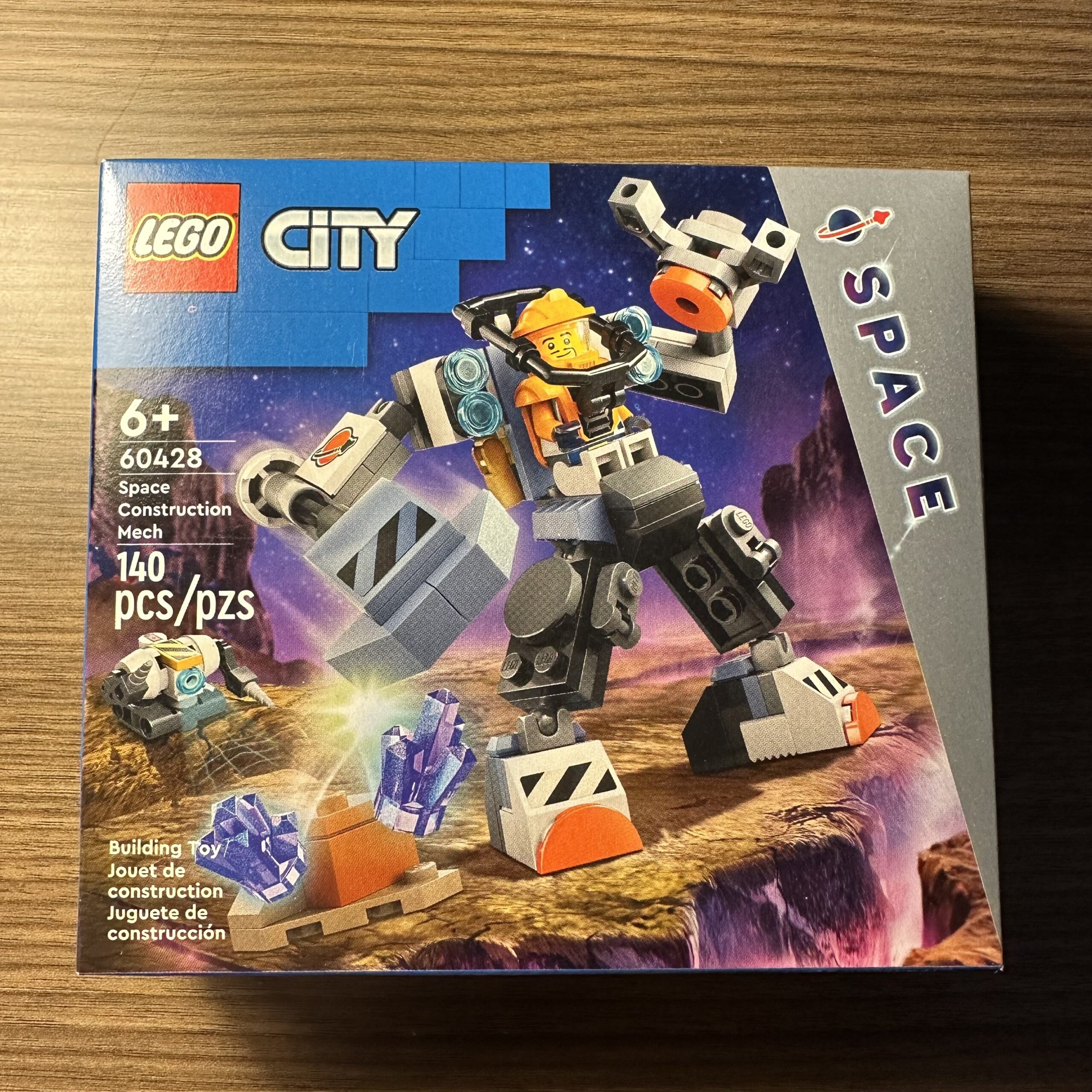 Box for LEGO City Space set 60428 Space Construction Mech. Box depicts an orange astronaut in a gray mech pounding some purple crystals off of some rocks. A small robot with two drill arms is in the background.