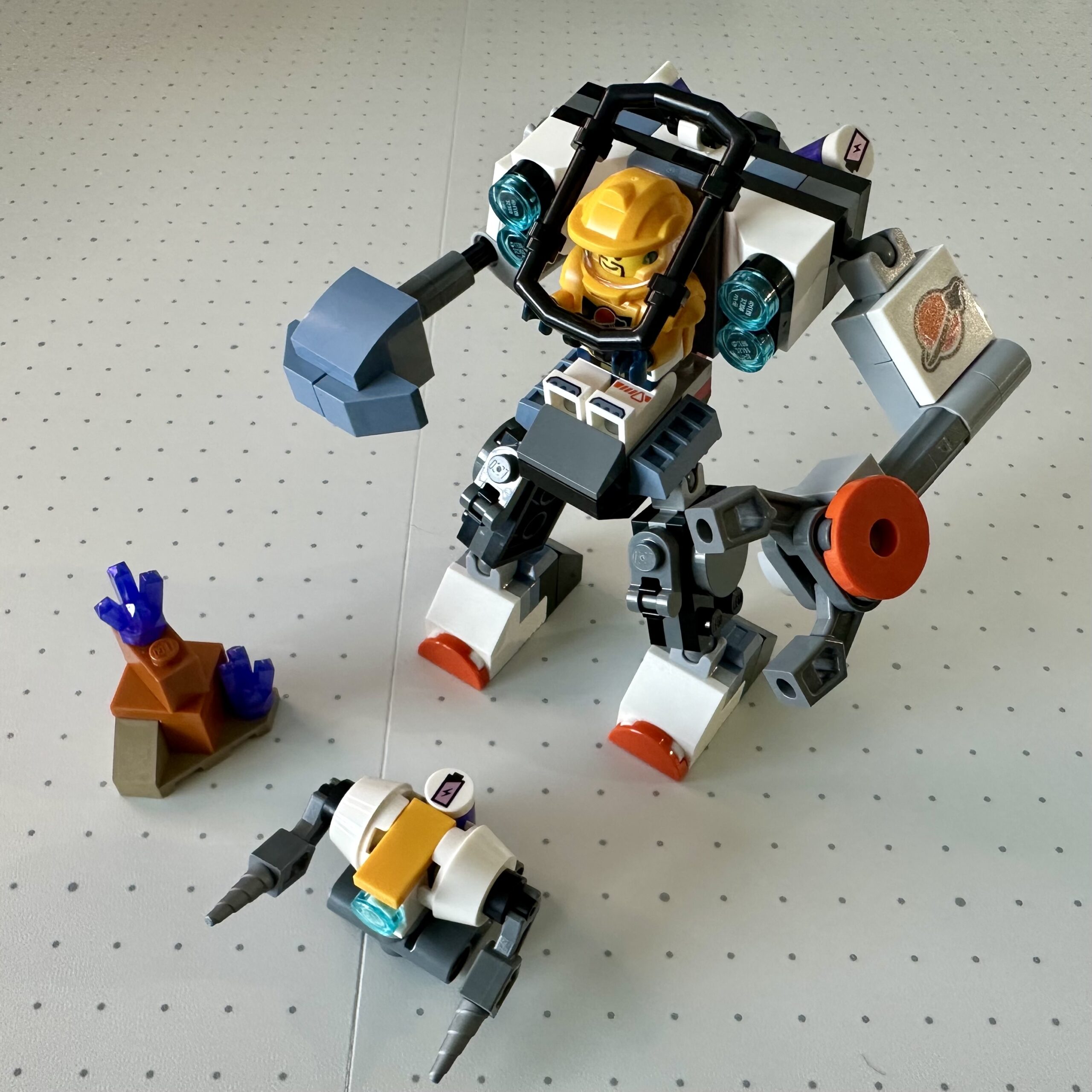 A gray and white LEGO mech in white and gray towers over a small robot with two drill arms and a small dark orange section of alien terrain with two purple crystals.