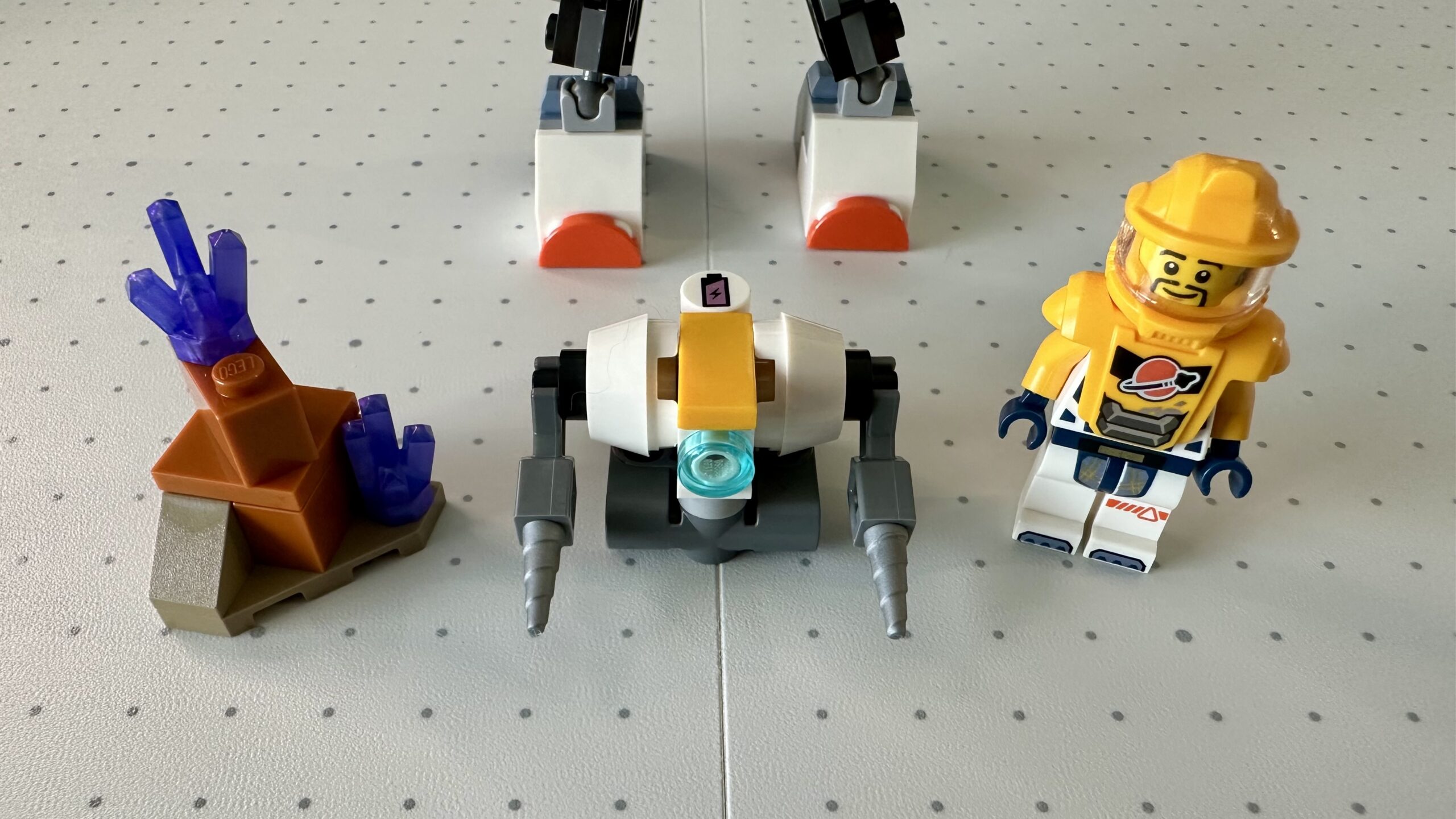 Close up of some LEGO. From left to right: a small section of alien terrain with purple crystals, a small mining robot with drill arms, and an astronaut in yellowish-orange armor.